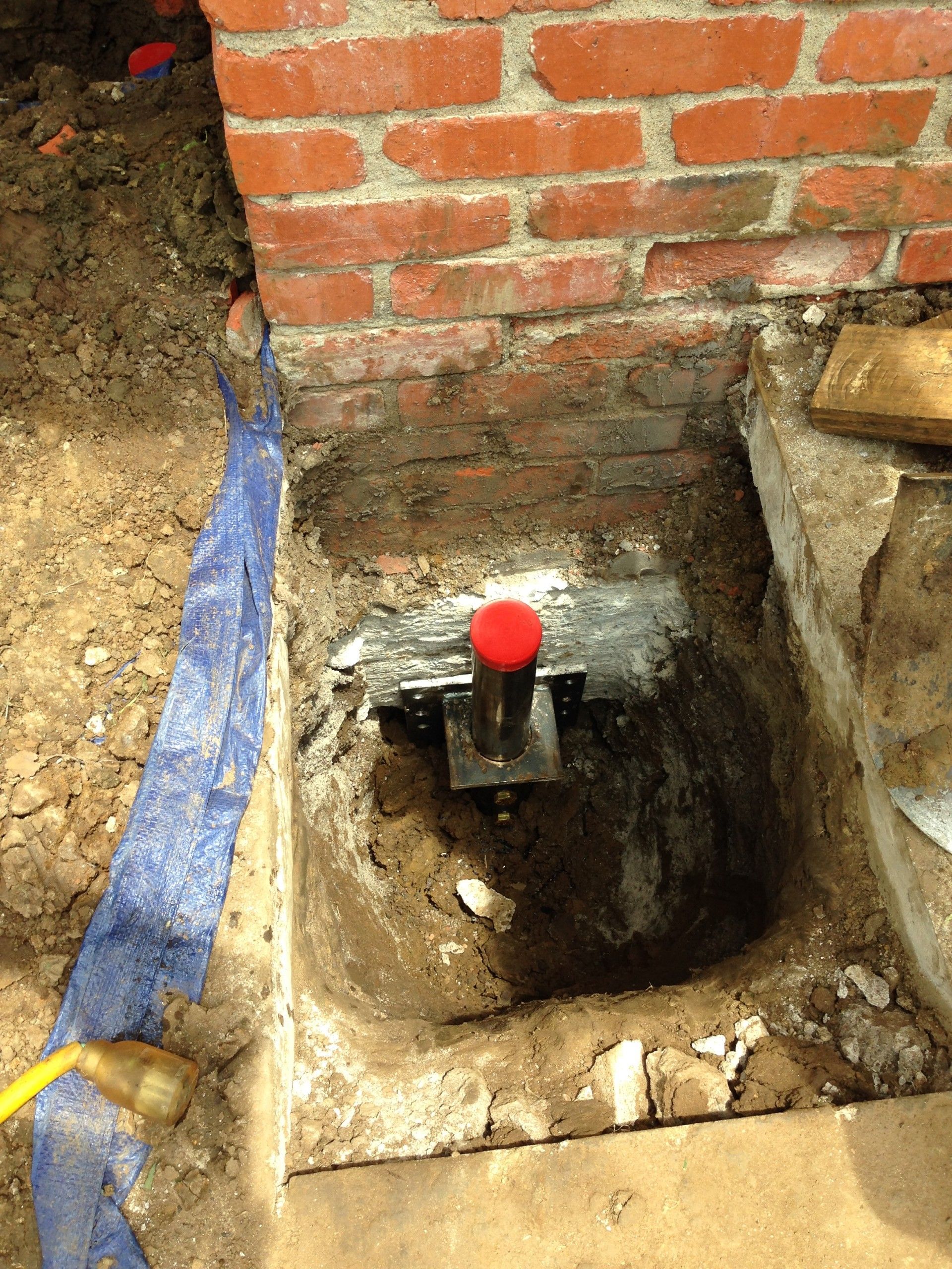 A metal pier support installed in a hole next to a brick wall foundation, featuring a prominent red cap.