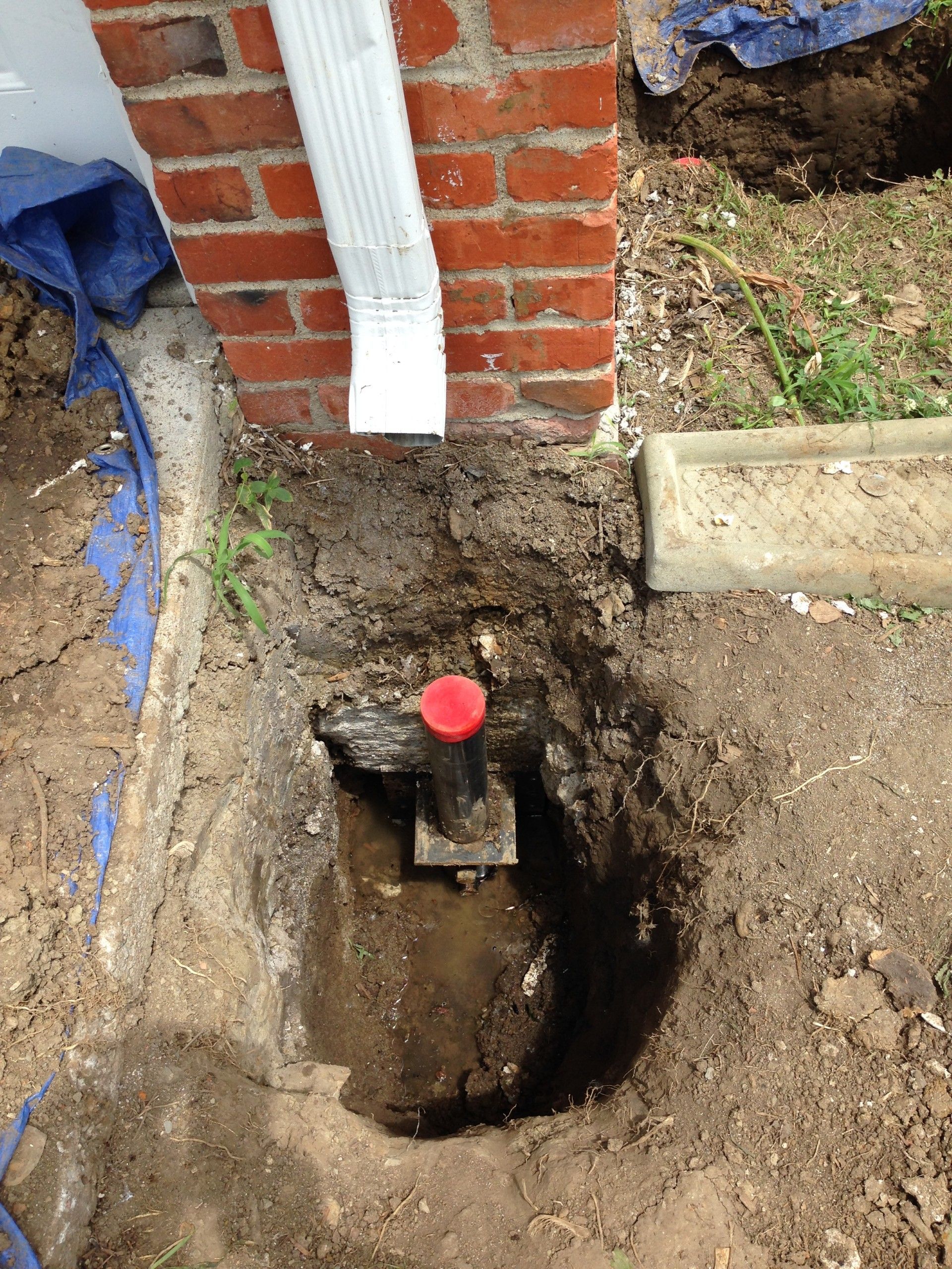 A vertical pipe with a red cap sits in a hole in the ground next to a red brick house wall and a white downspout.