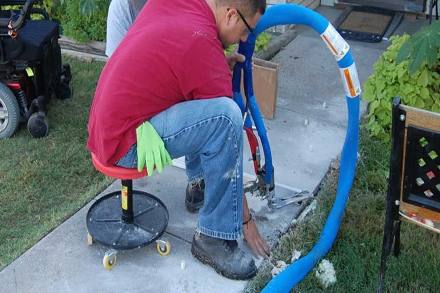 A person in a red shirt sits on a rolling stool, using equipment to inject foam into cracks in a concrete walkway.