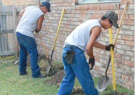 Two workers in gray shirts and jeans use shovels to dig alongside a brick wall.