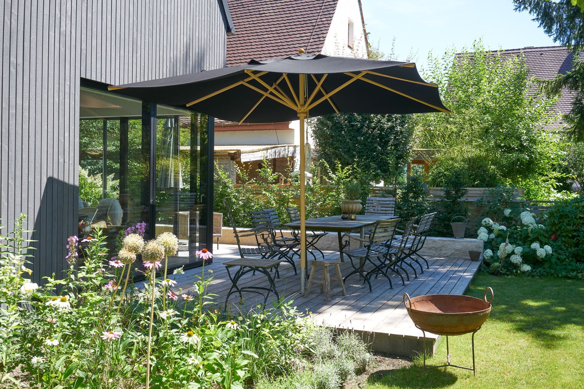 Garden patio with black umbrella, outdoor table and chairs, and flowers beside a house
