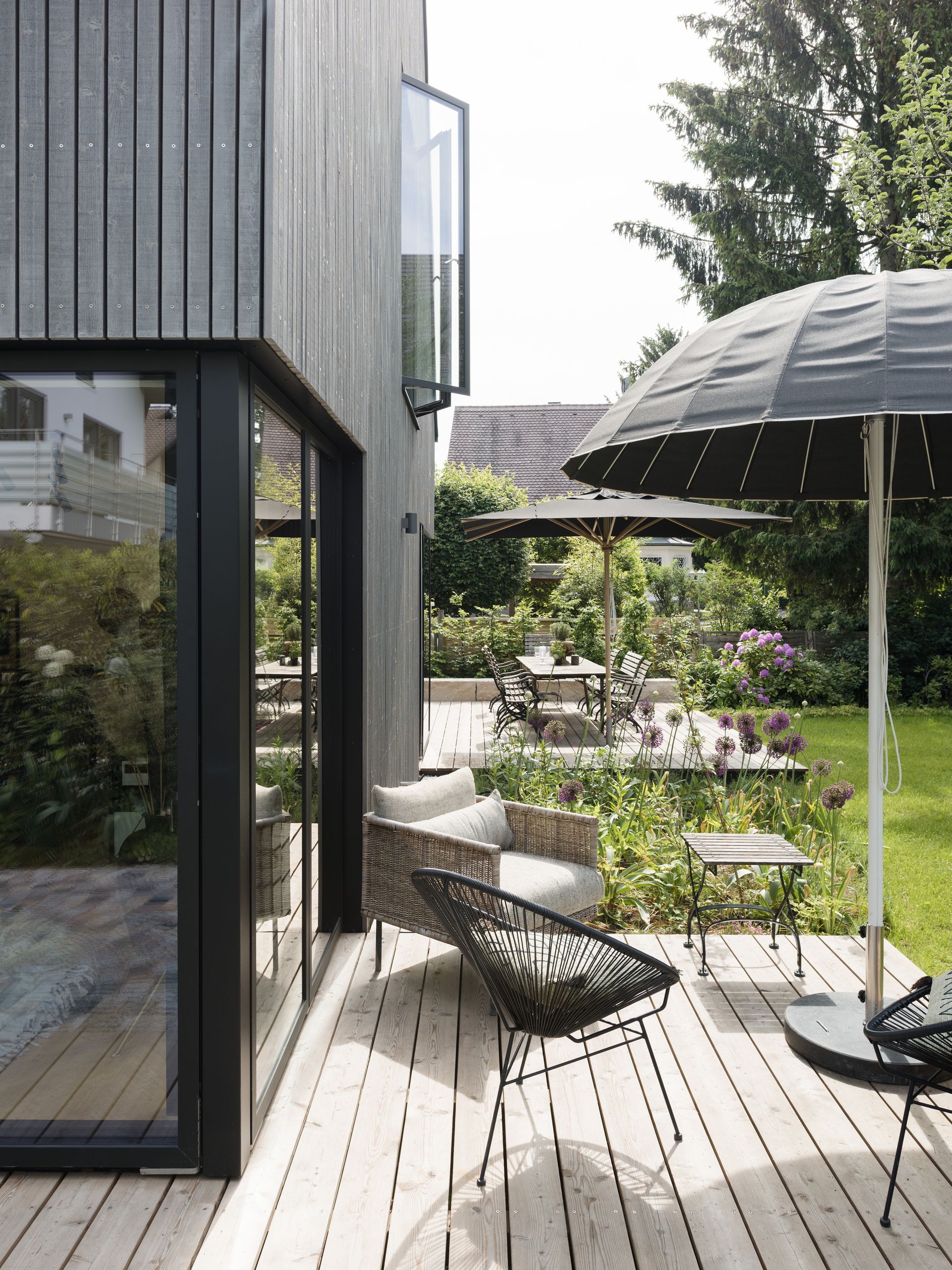 Modern patio beside a black house, with wicker chair, umbrella, steps, and lush garden greenery