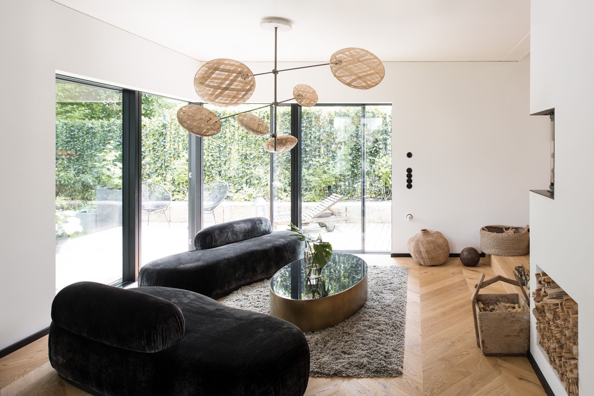 Modern living room with black velvet sofas, woven pendant lights, and large glass doors opening to a patio.