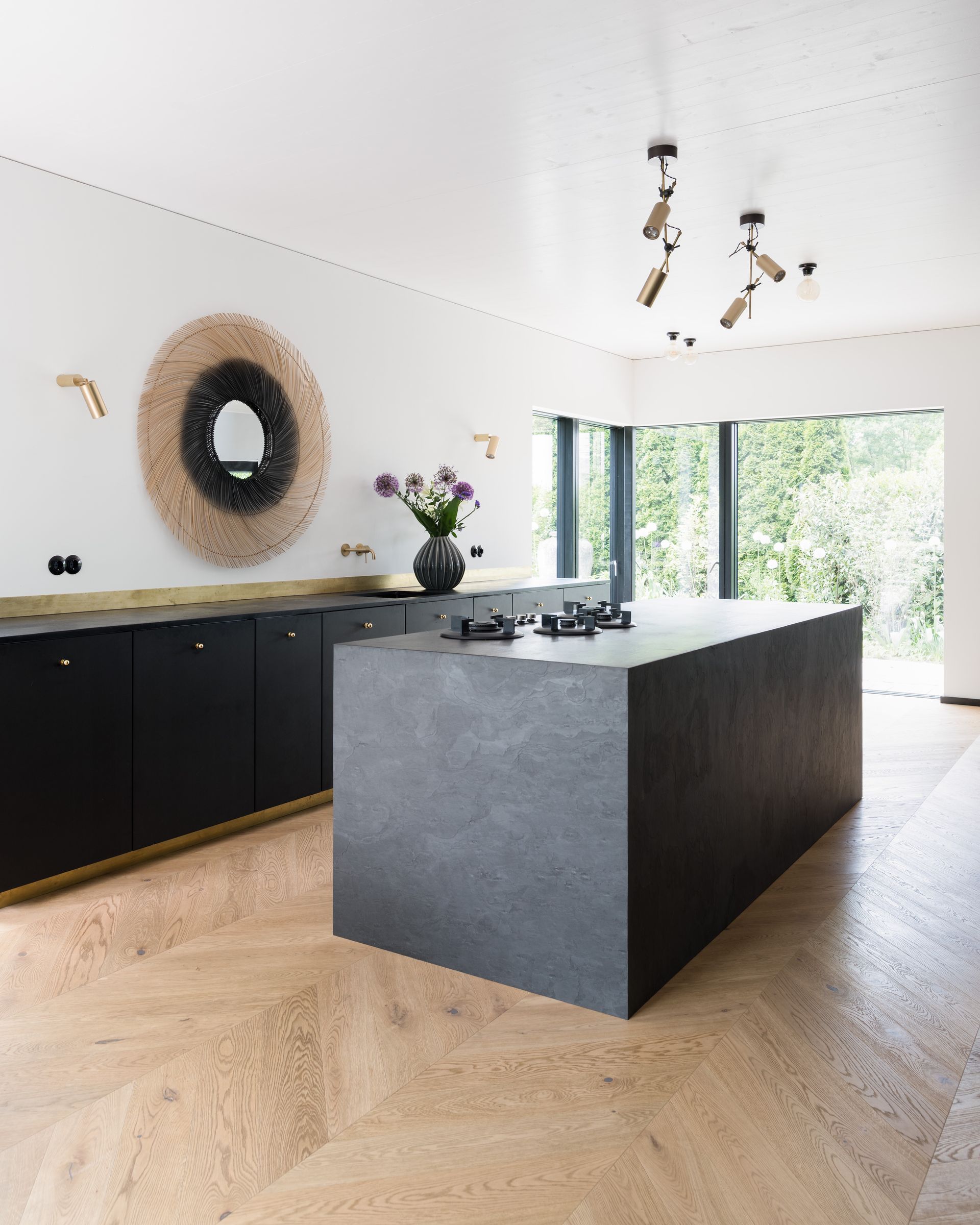 Modern kitchen with black cabinetry, large gray island, wood floor, and bright glass doors overlooking greenery