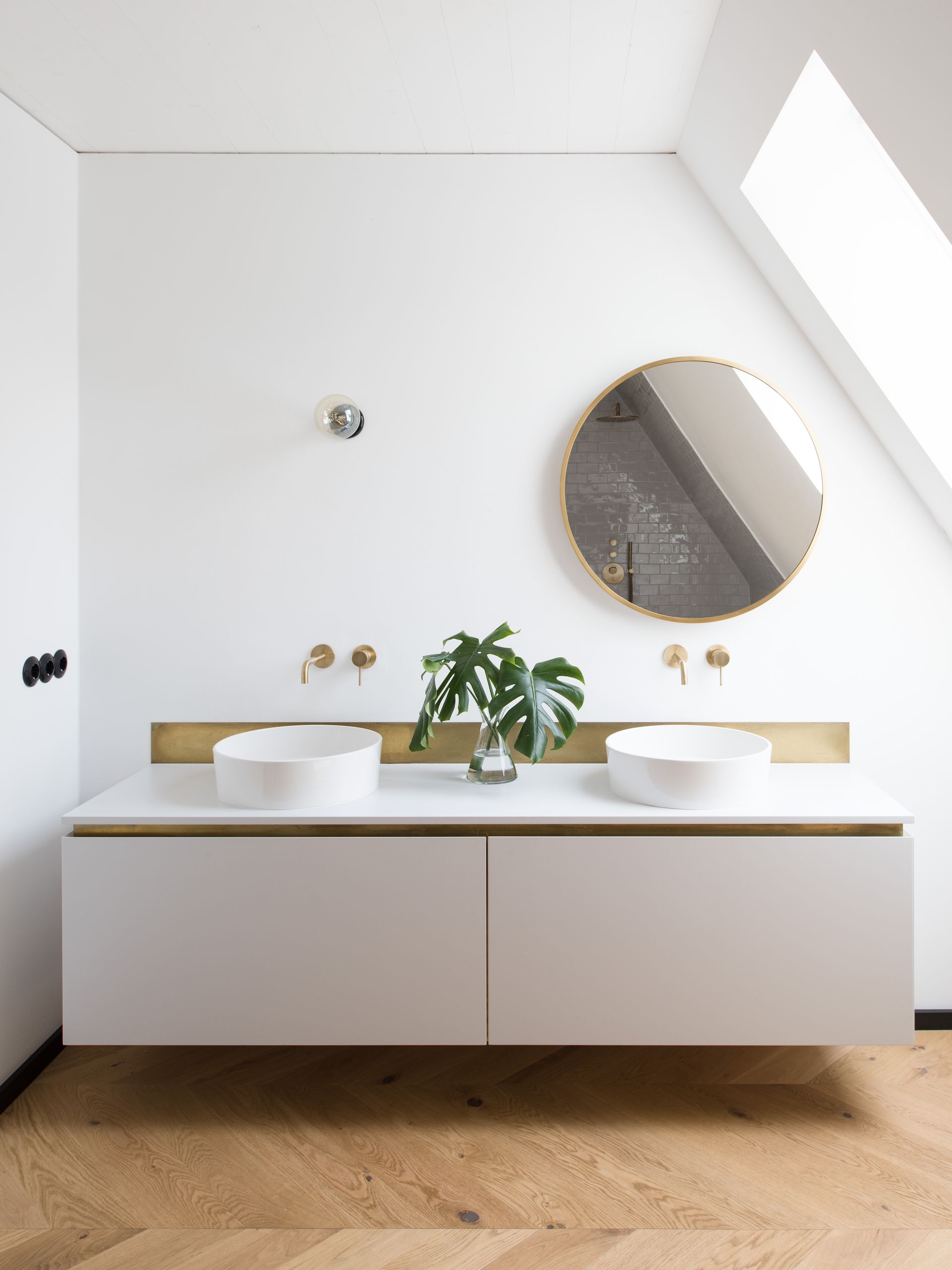 Modern white bathroom vanity with twin sinks, gold fixtures, round mirror, and wood floor under skylight.