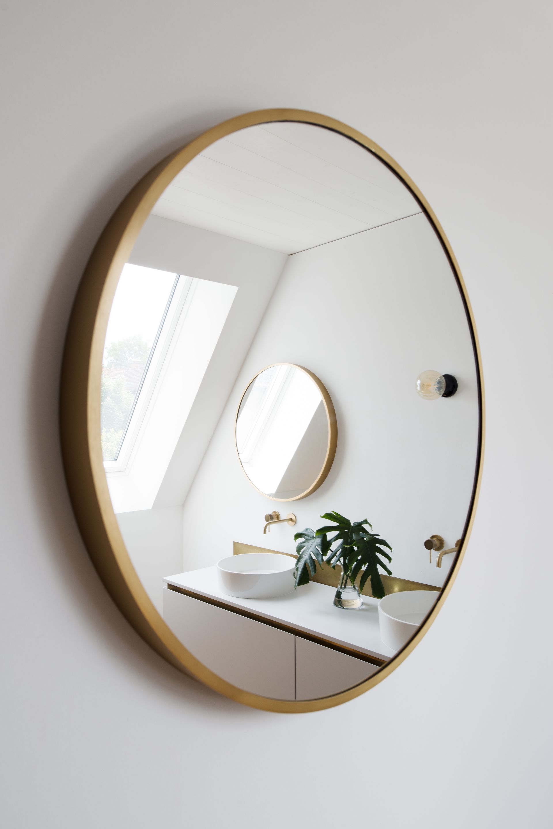 Round gold-framed mirror reflecting a bright bathroom vanity with sink, plant, and skylight