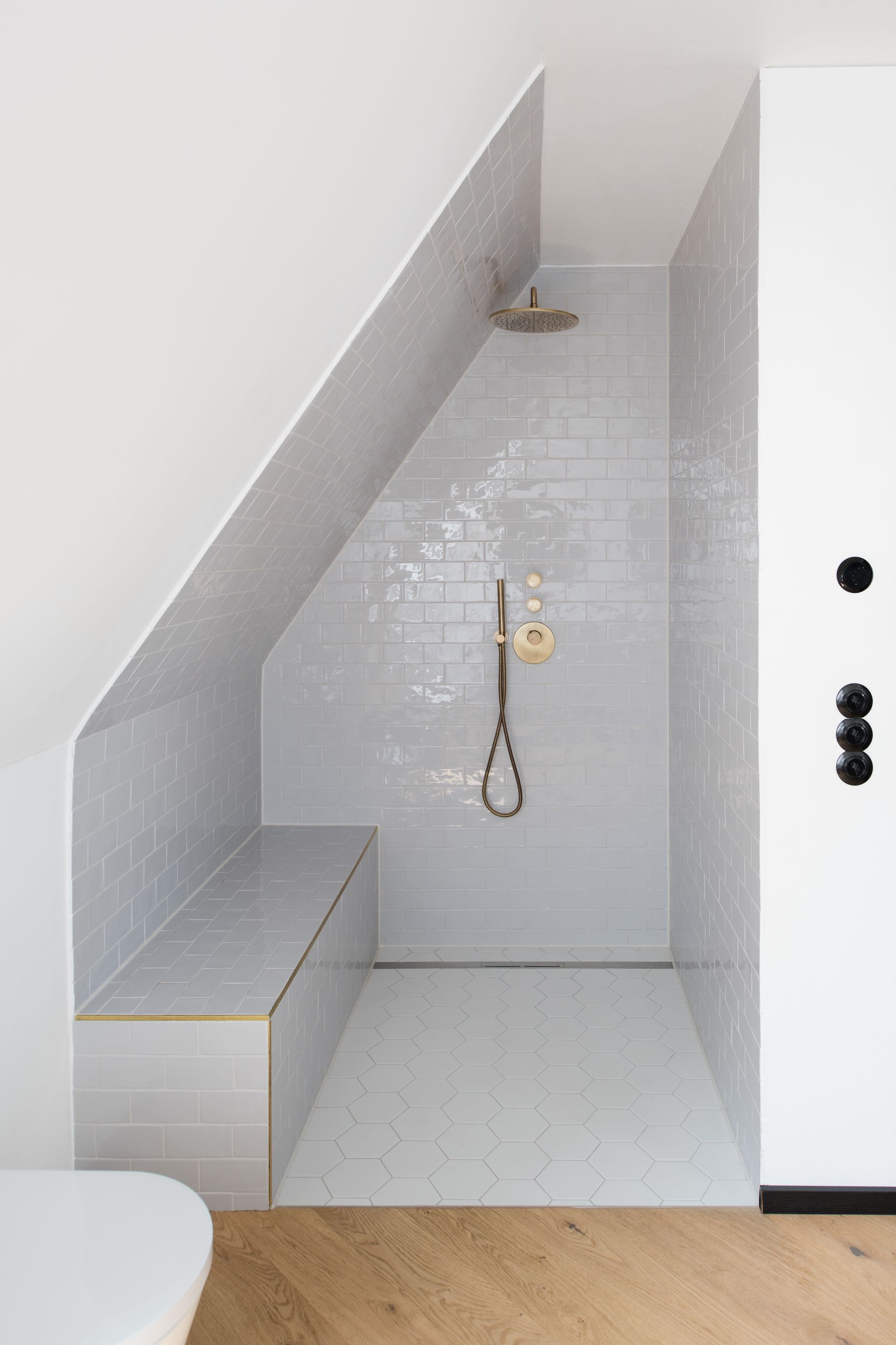 Modern white tiled shower under a sloped ceiling with built-in bench and showerhead.