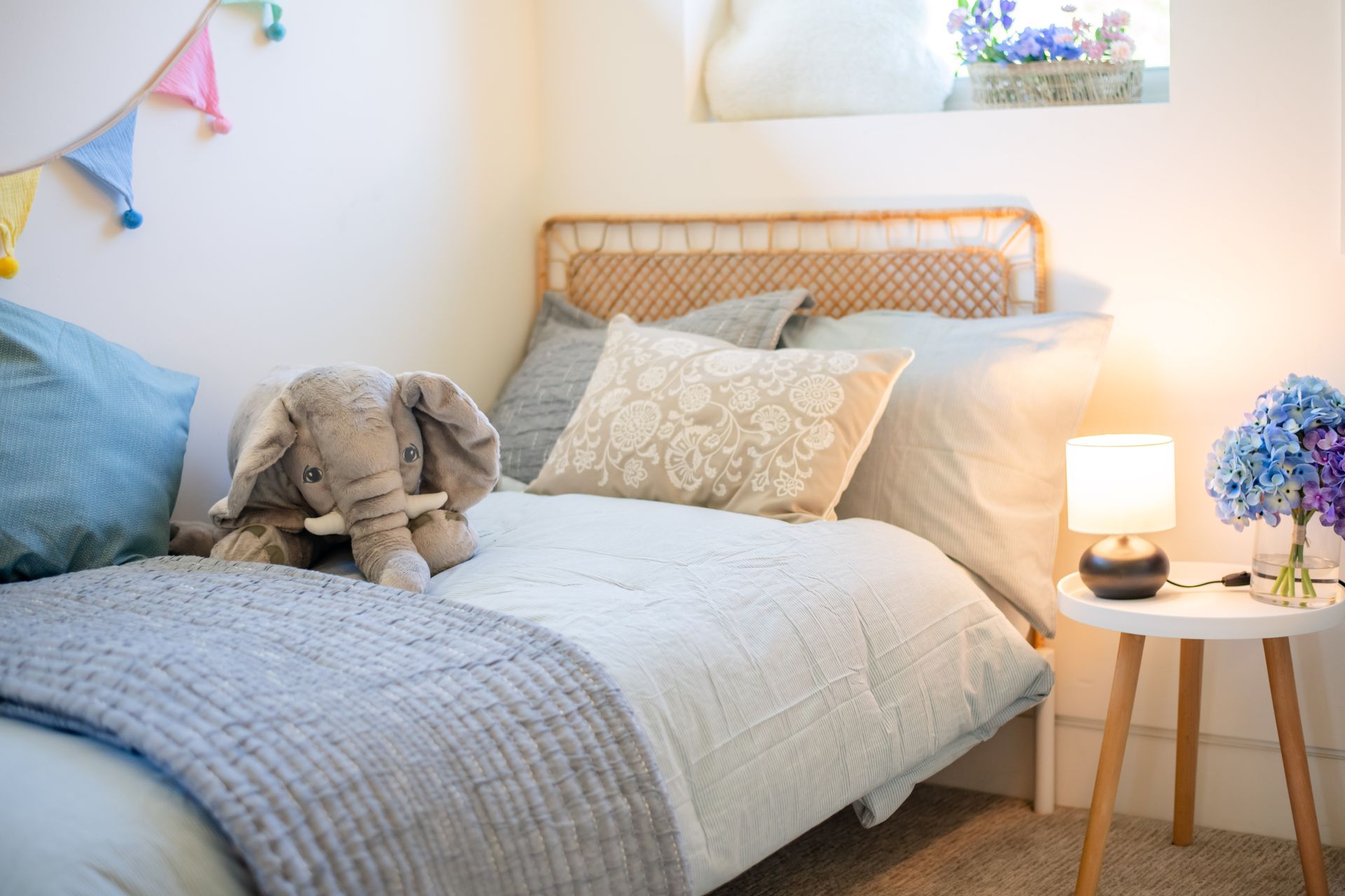 Cozy bedroom with a white bed, gray blanket, stuffed elephant, bedside lamp, and blue flowers on a small table