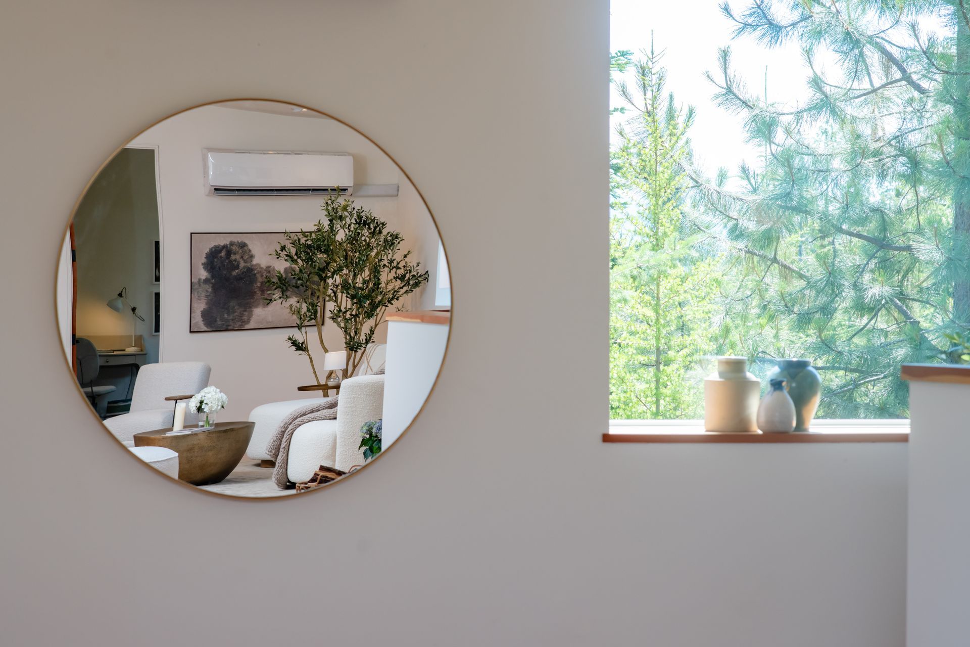 Round mirror reflecting a cozy living room beside a bright window with potted plants