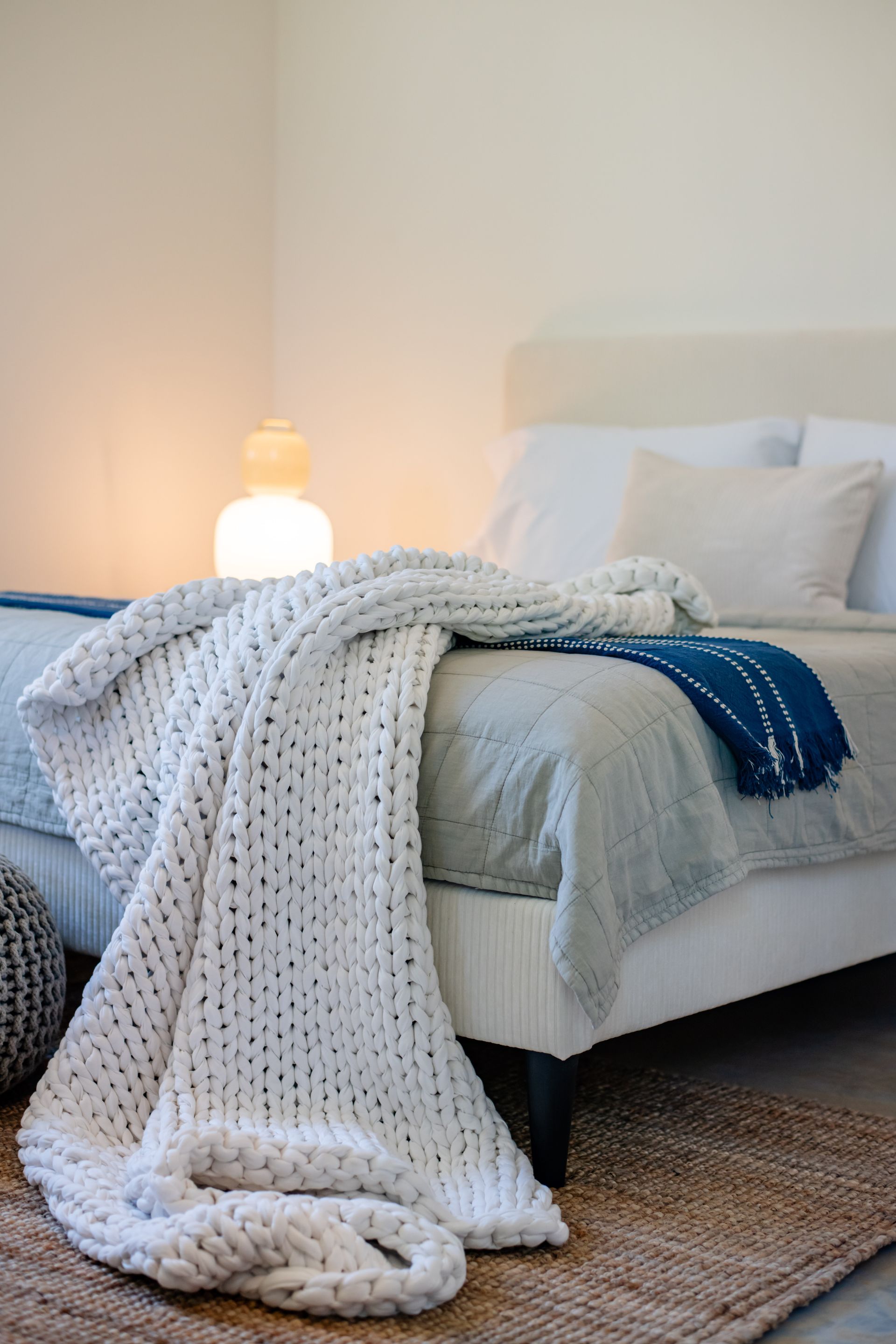 Cozy bedroom with a neatly made bed, draped white knit blanket, blue throw, and warm bedside lamp