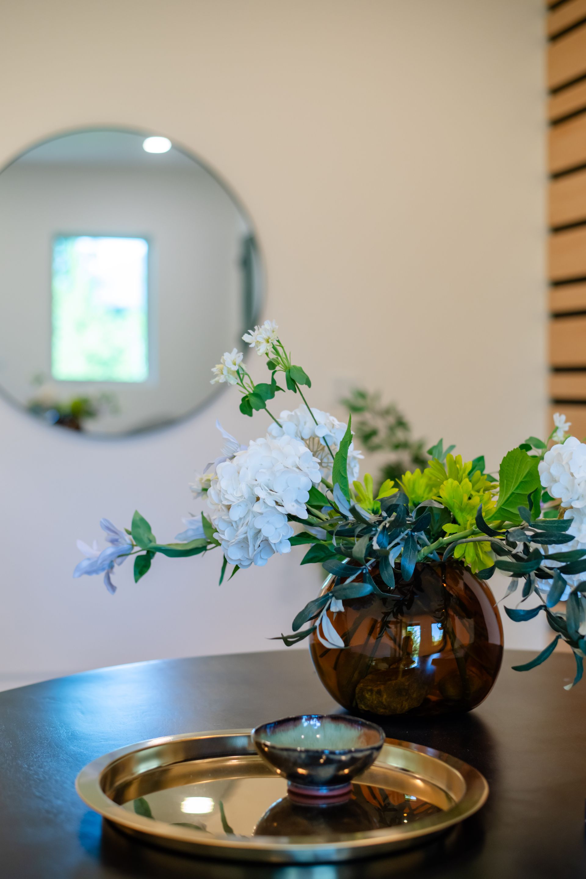 Floral arrangement in a brown vase on a table, with a mirrored tray in the foreground and a round wall mirror behind.