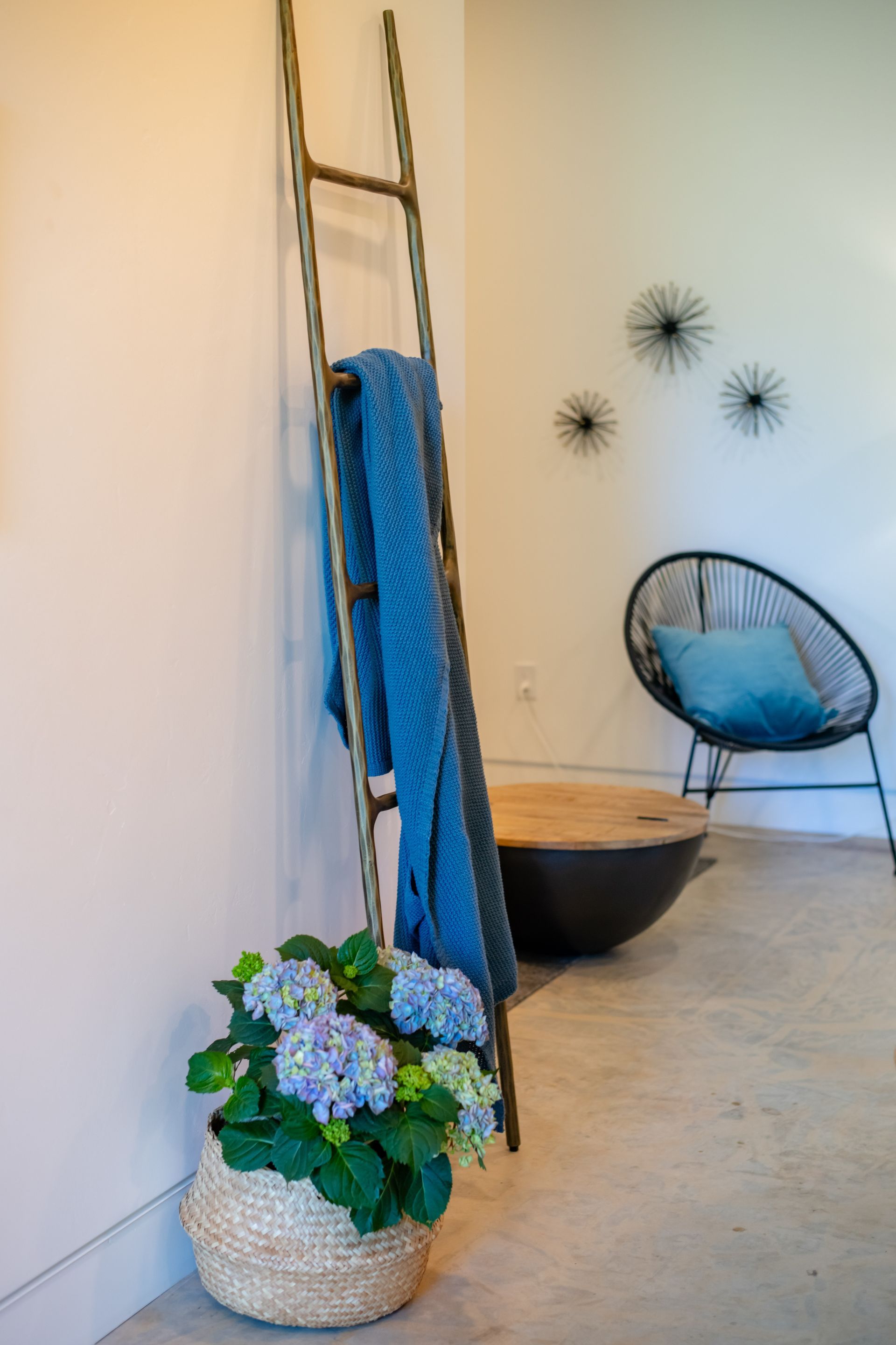 Cozy corner with a blue chair, ladder, black bowl, wall decor, and a flower basket on a concrete floor