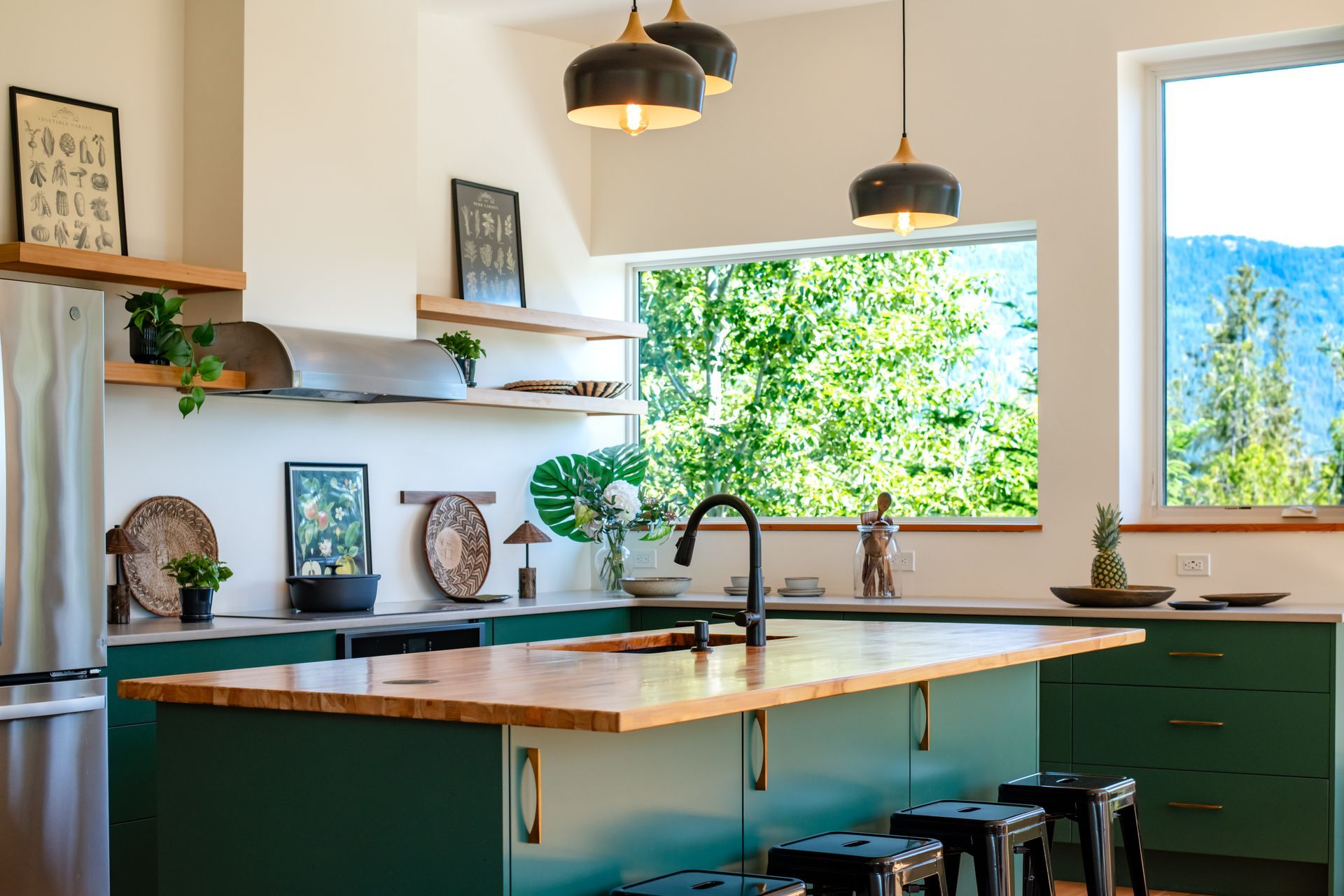Modern kitchen with green cabinets, wooden island, black fixtures, and large windows overlooking greenery