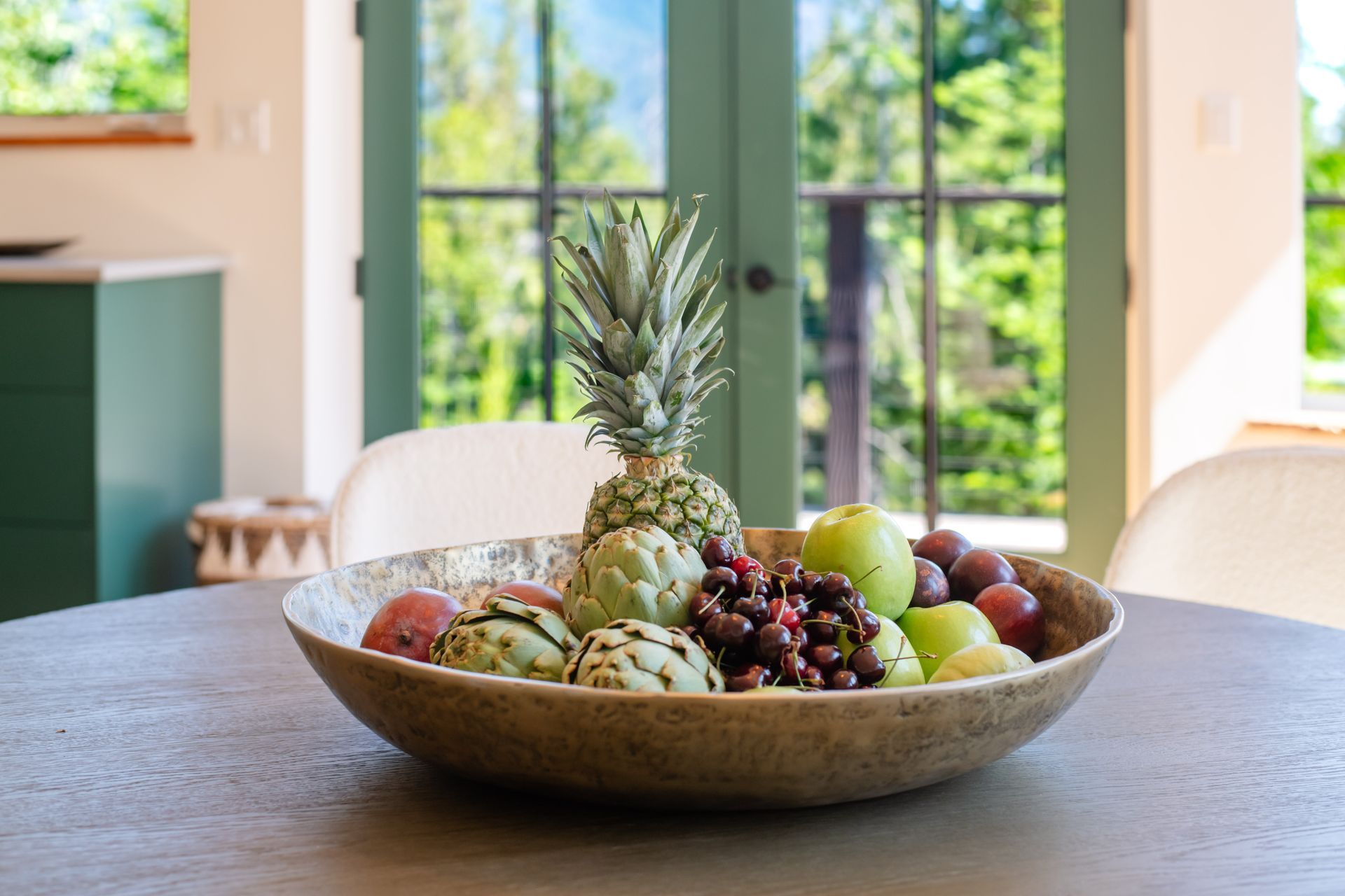 Fruit bowl centerpiece on a wooden table in a bright room with large windows and greenery outside