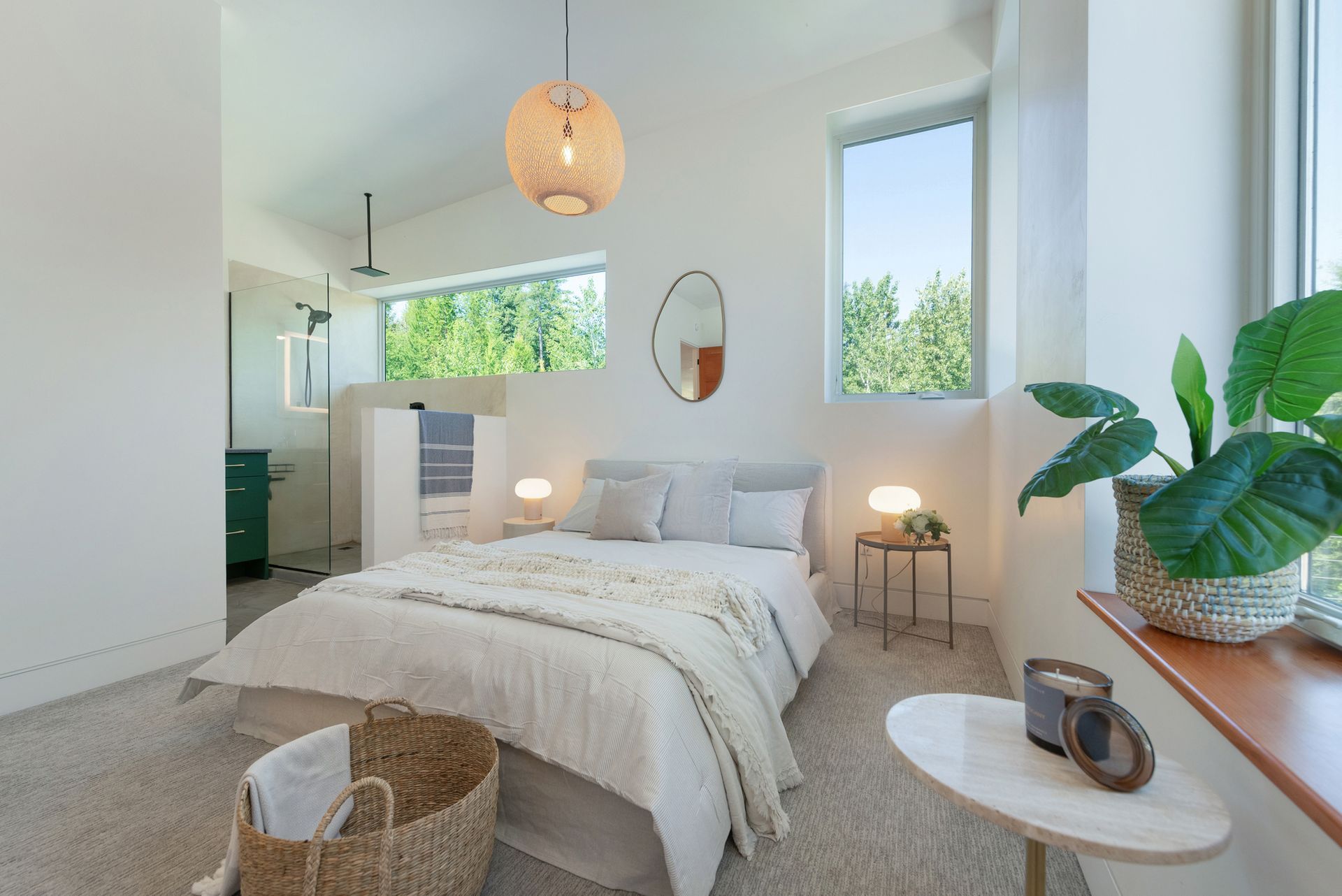 Bright modern bedroom with white bed, large windows, woven lamp, plants, and cozy neutral decor.