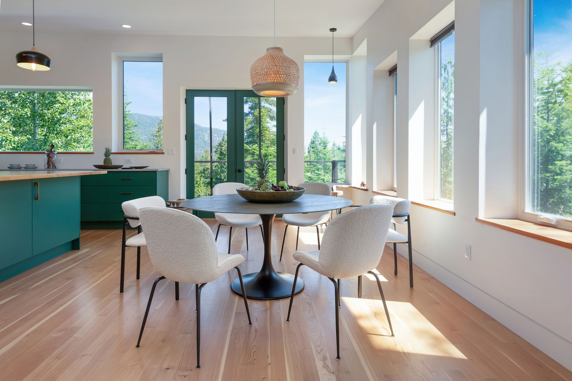 Bright dining area with round table, white chairs, teal kitchen cabinets, and sunlight through large windows