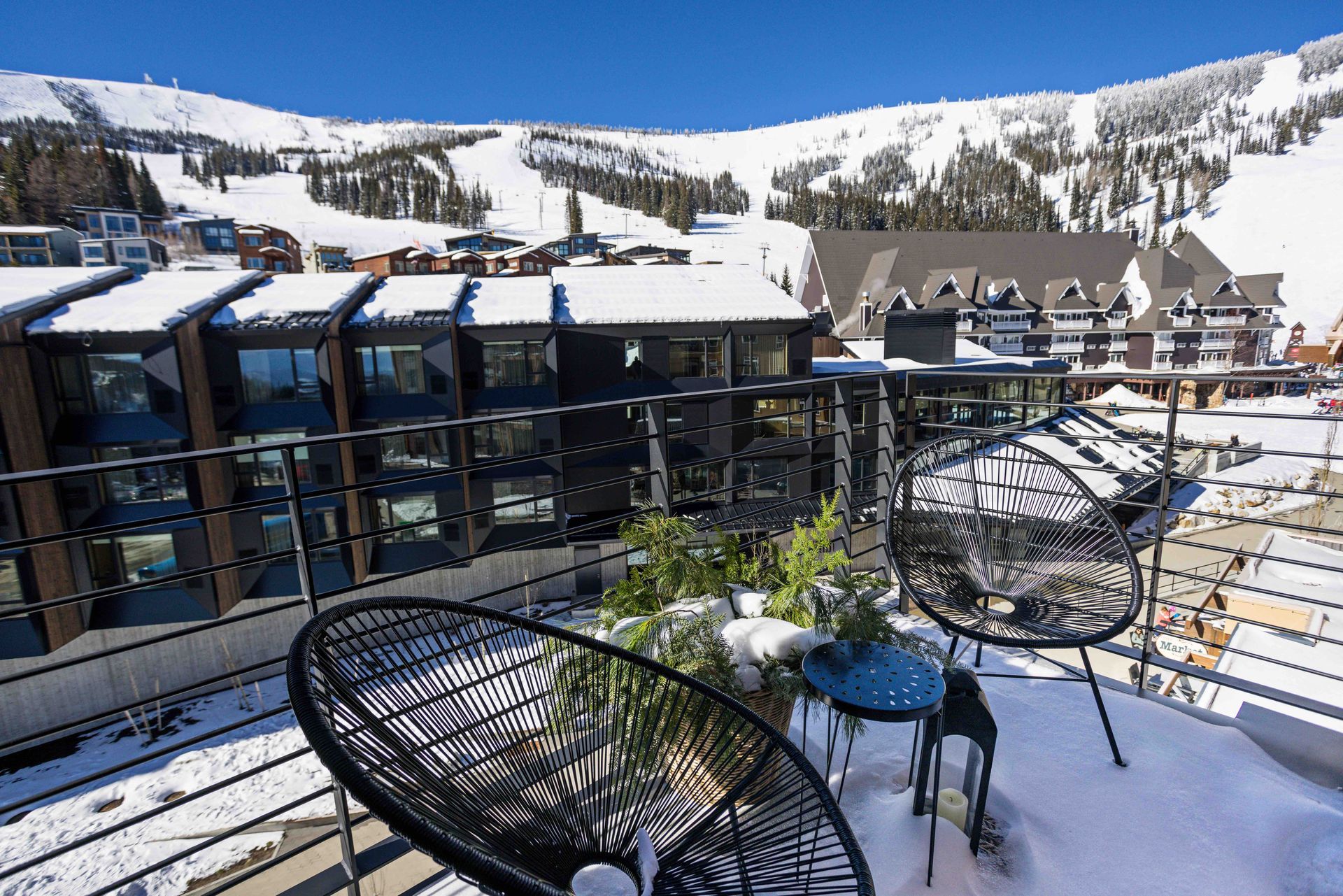 Snowy resort terrace with black chairs, blue tables, and mountain lodge buildings in the background