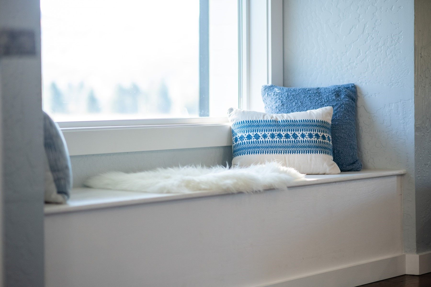 White window seat with fluffy cushion and blue patterned pillows beside a bright window