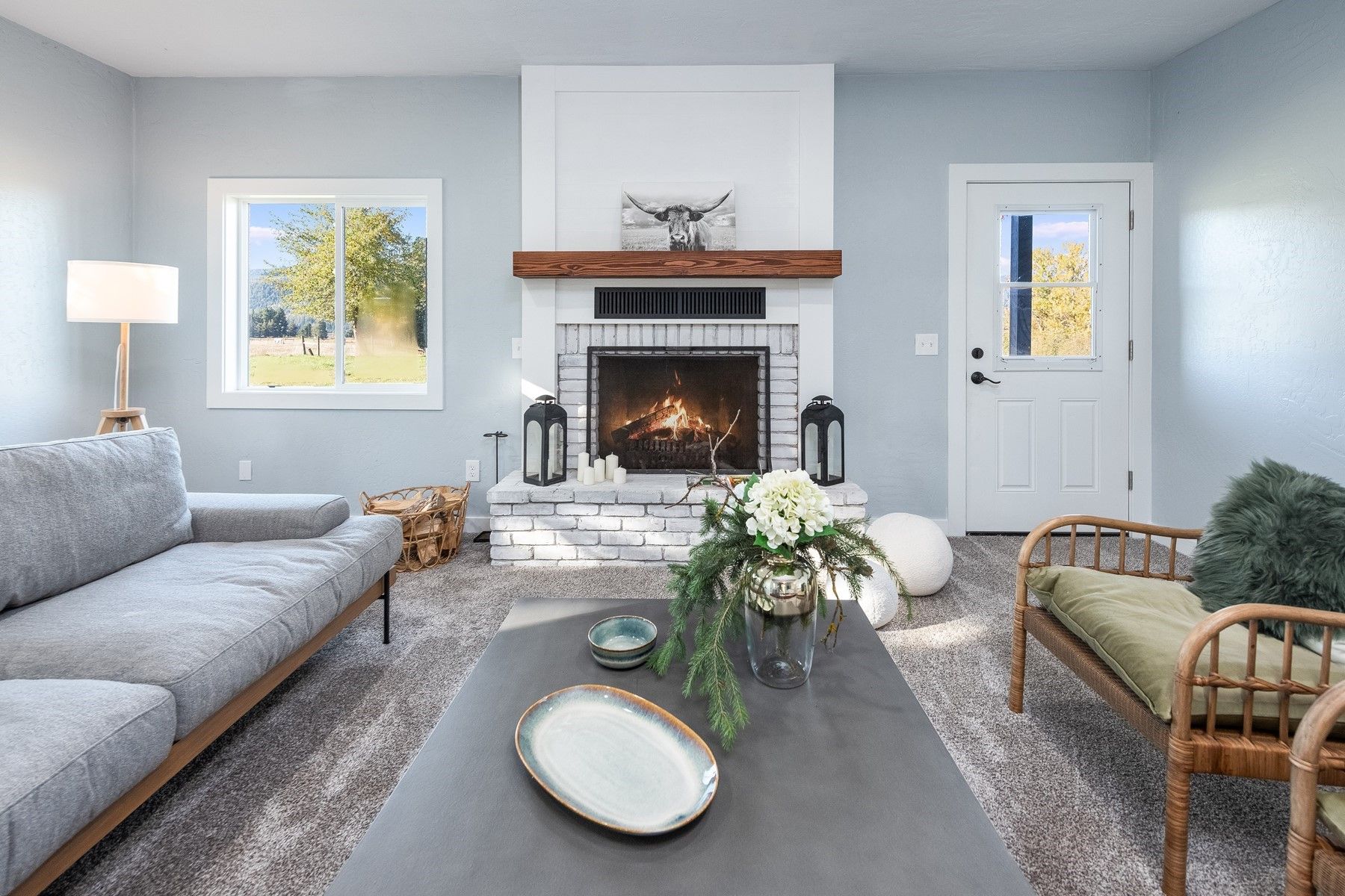 Bright living room with gray sofa, wicker chair, coffee table, fireplace, and large windows