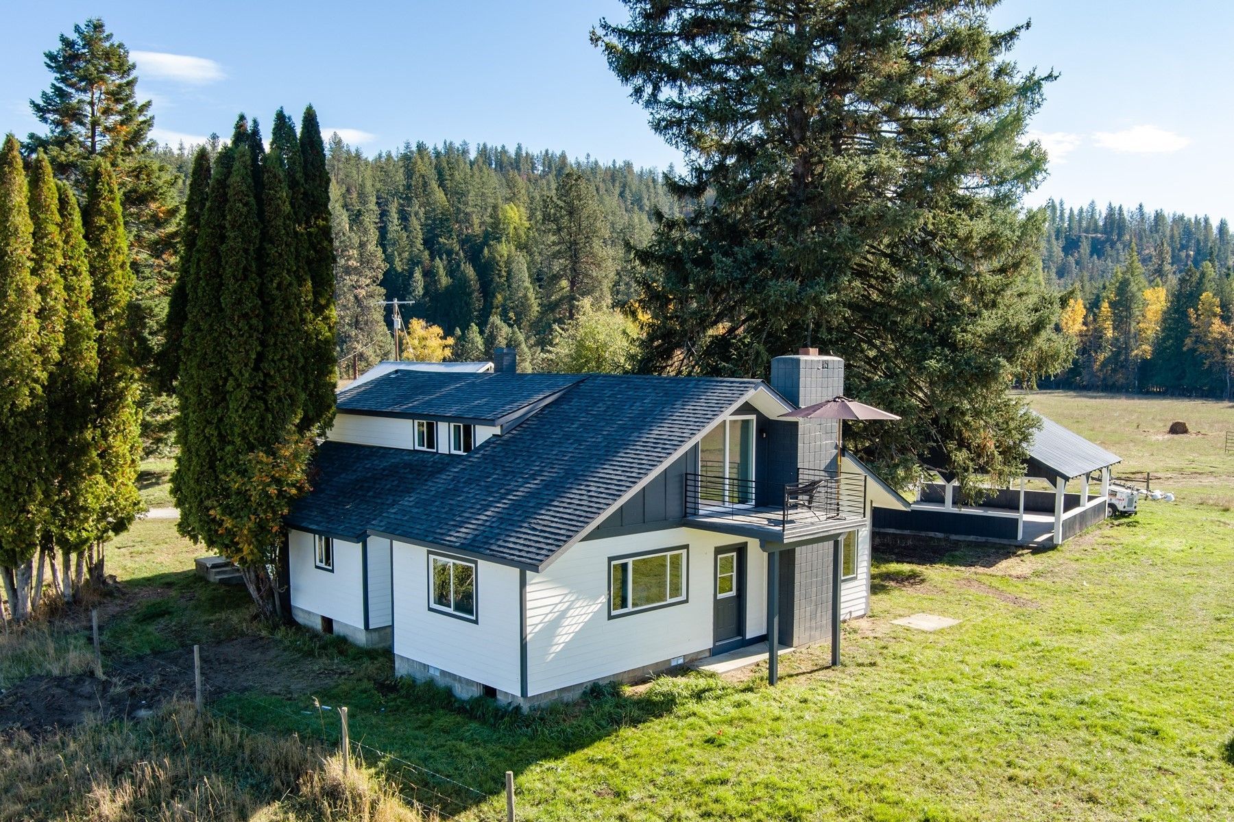 White house with dark roof in a grassy wooded hillside clearing, with tall trees and mountains in the background