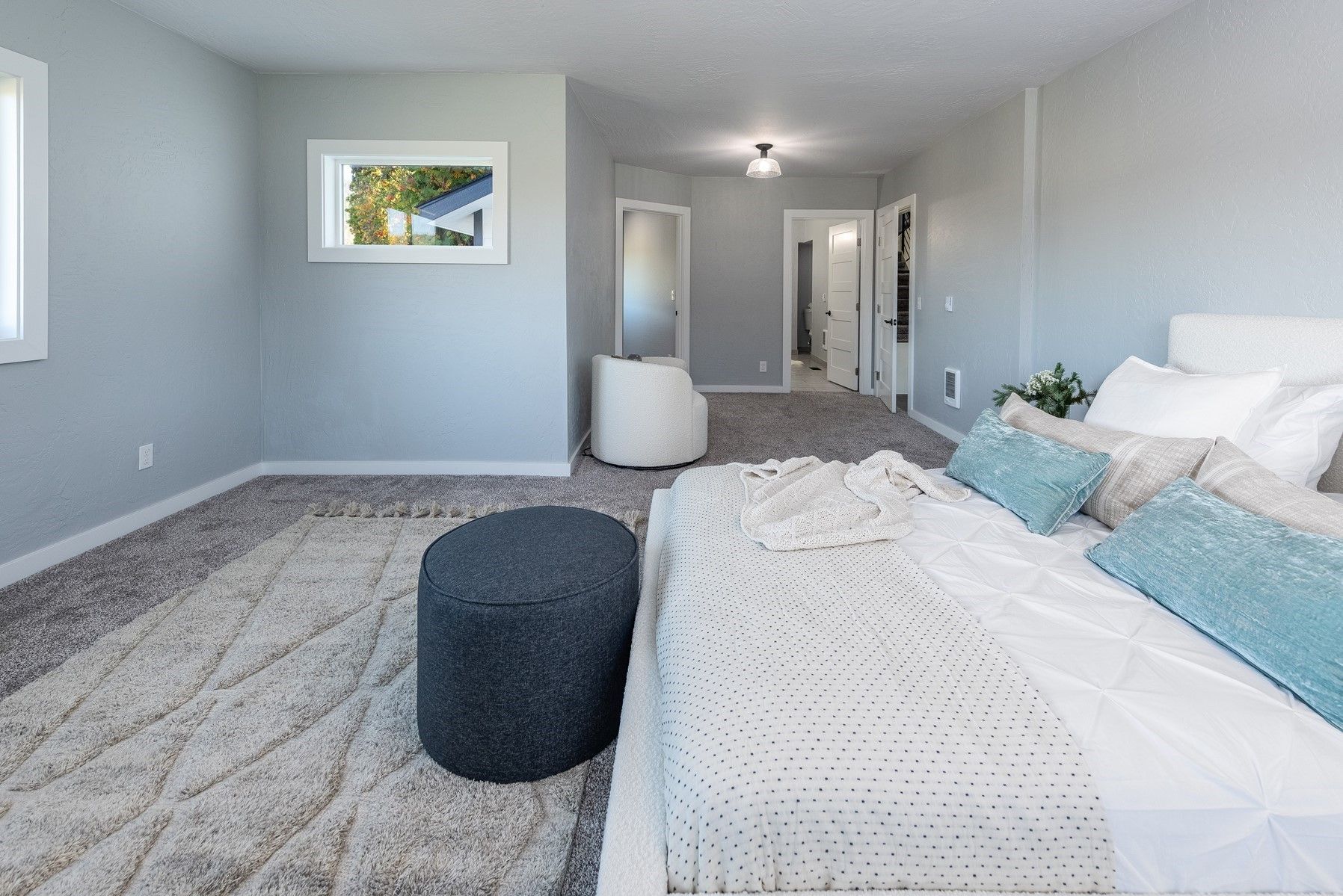 Bright bedroom with a bed, gray carpet, ottoman, wall art, and open doorway to a hall