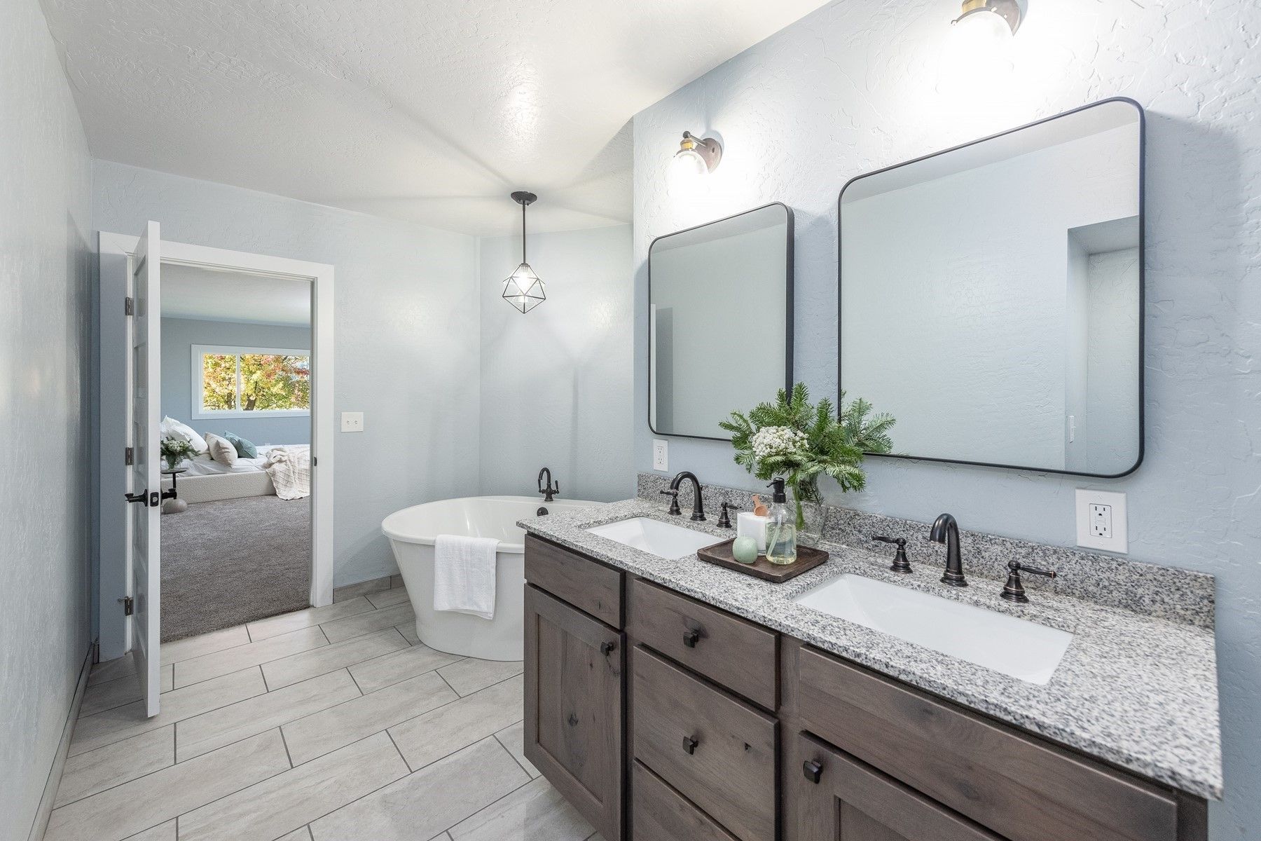 Bright bathroom with double vanity, mirrors, freestanding tub, and doorway to a bedroom.