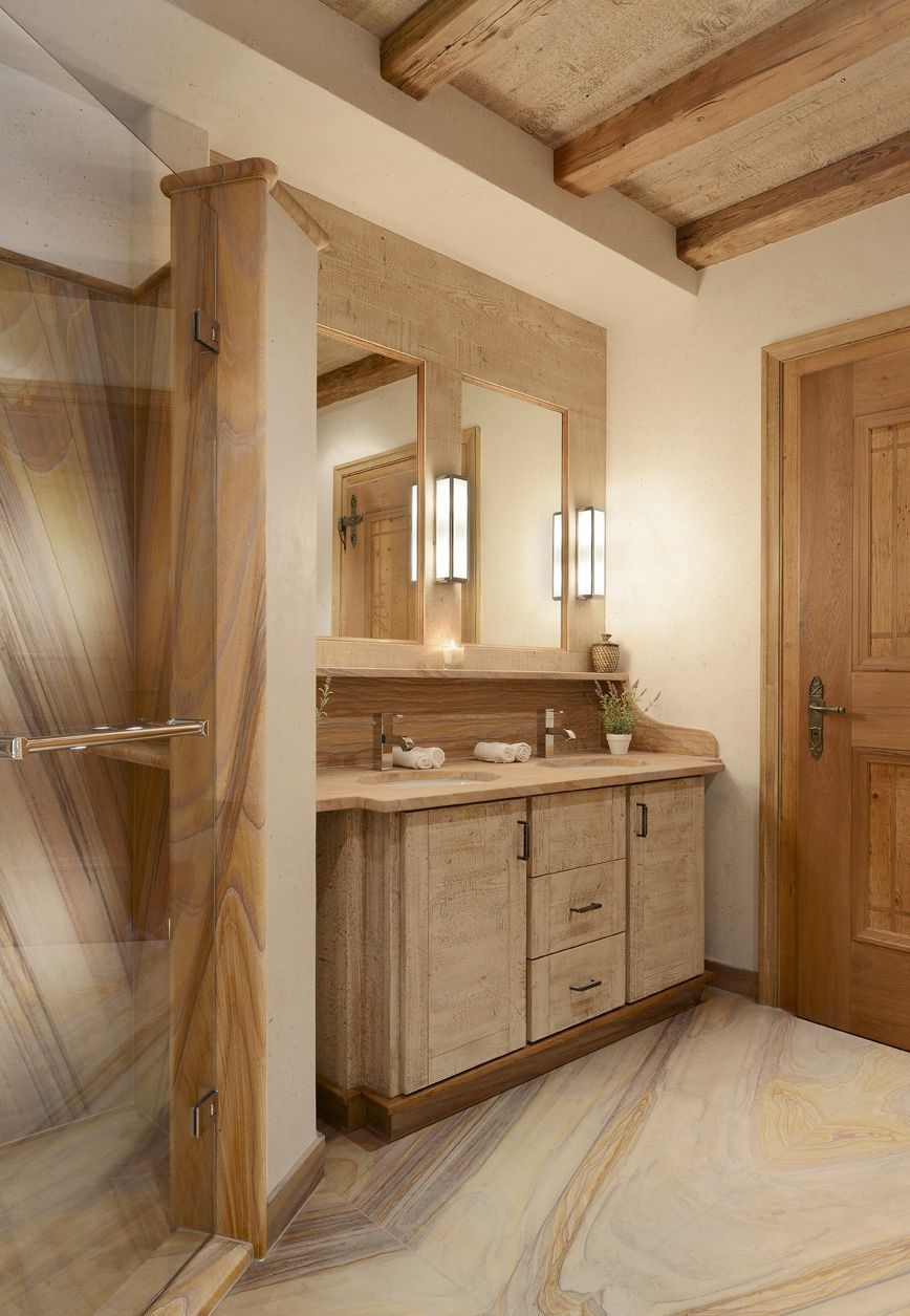 Rustic bathroom with wood vanity, mirror, sconces, and a wood door; beige stone walls and floor.