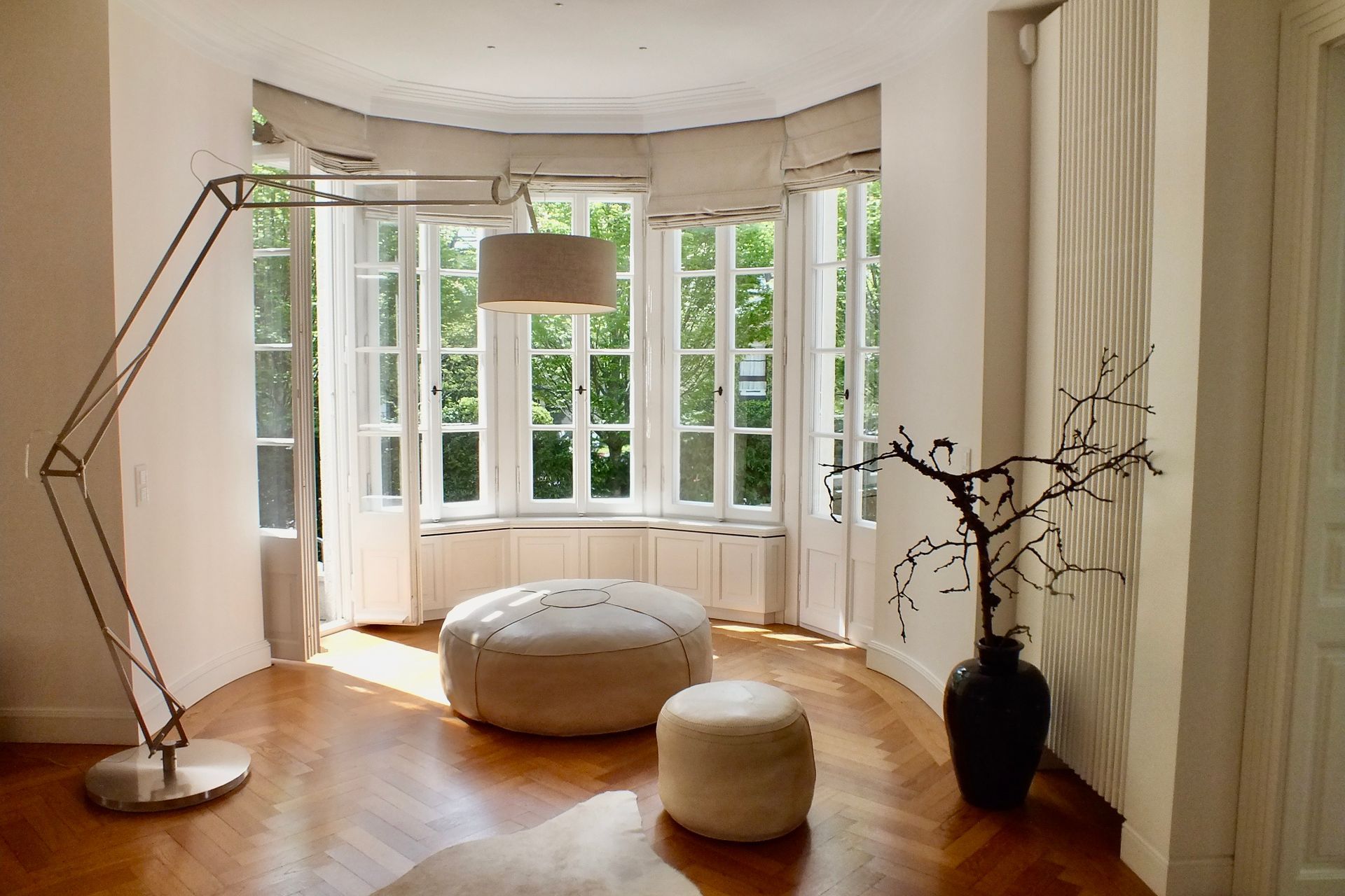 Bright living room with curved bay windows, hardwood floors, white ottoman, and floor lamps.