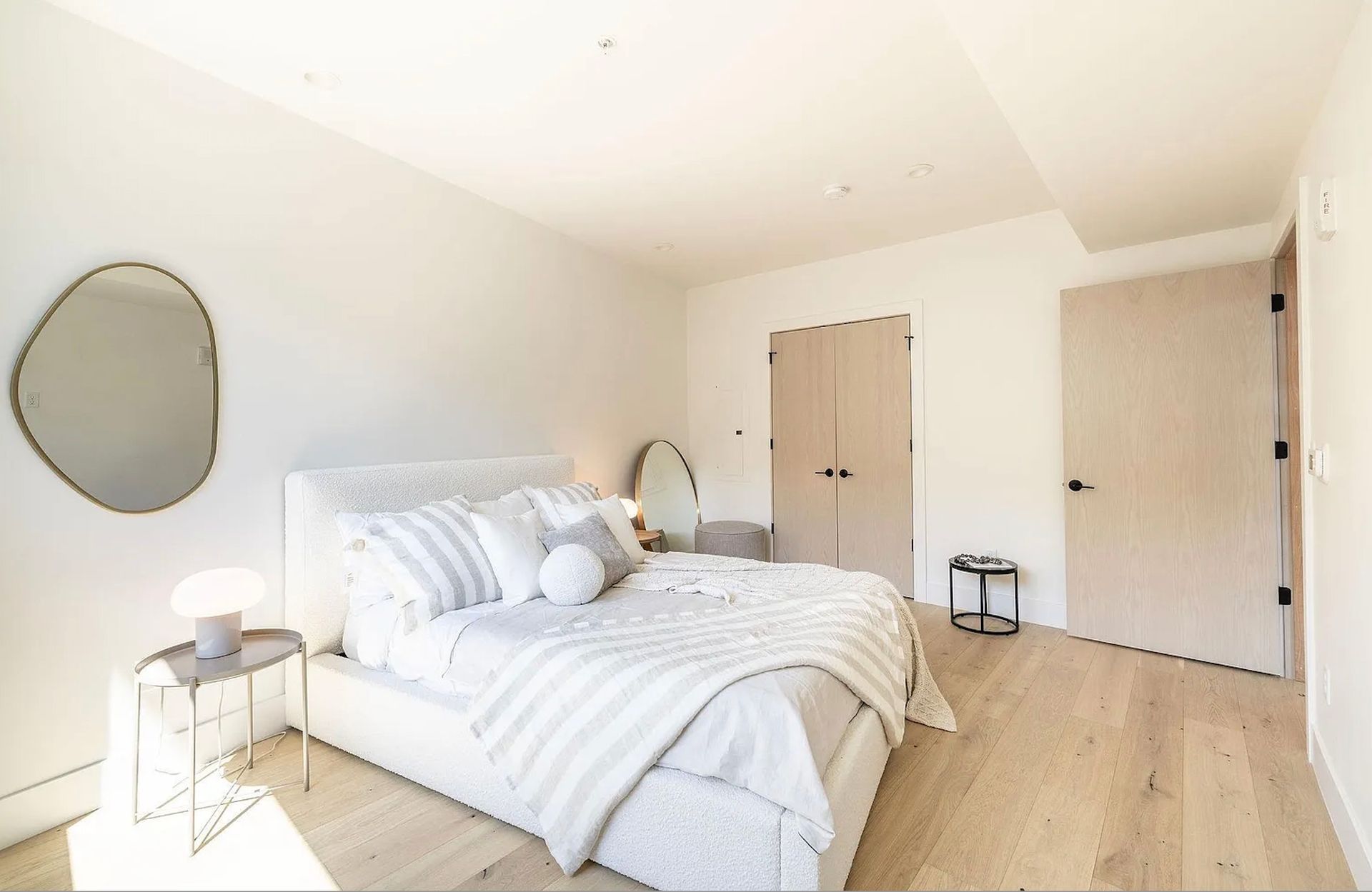 Bright minimalist bedroom with white bedding, round mirror, wood floors, and double doors.