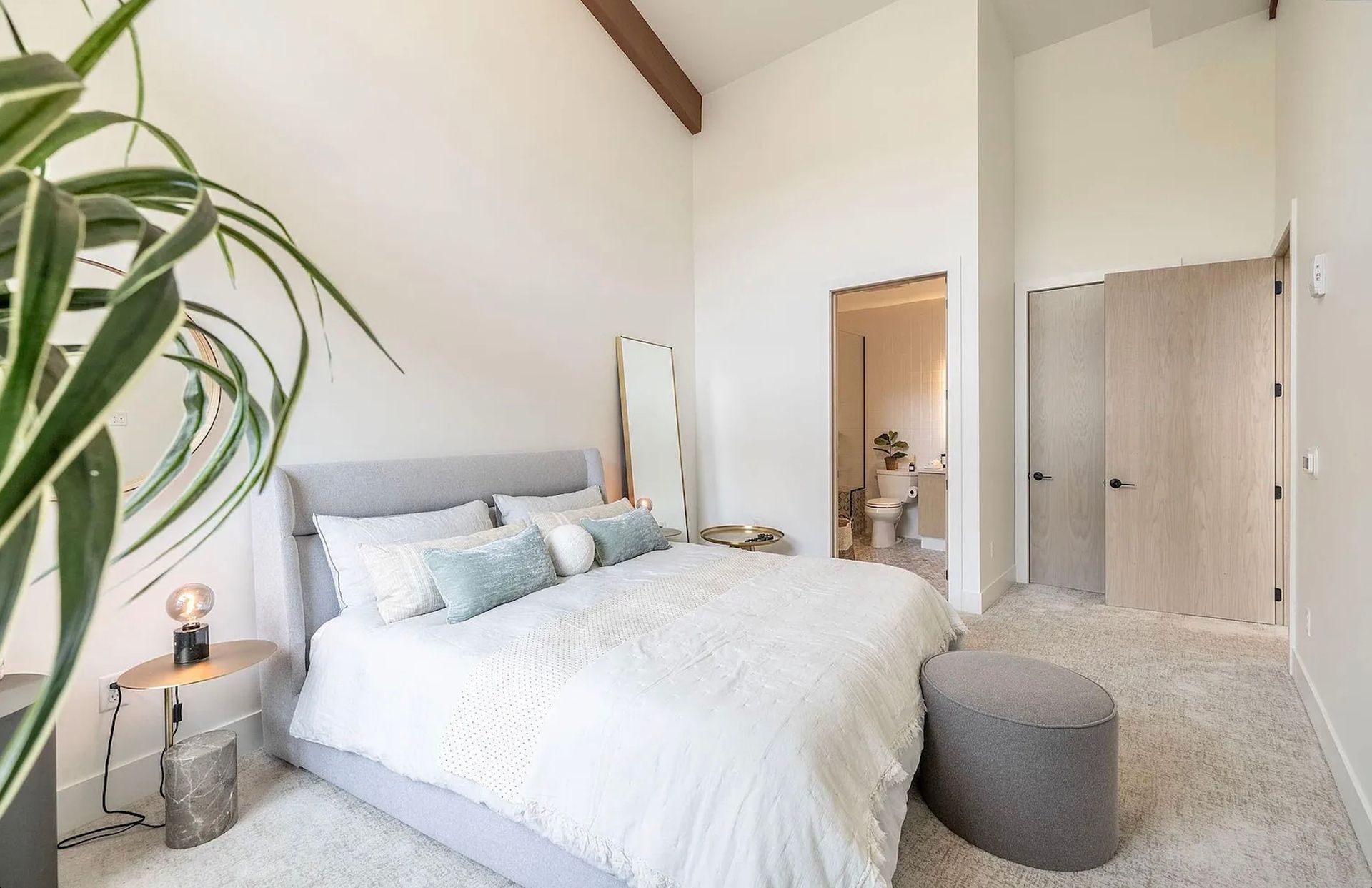 Bright bedroom with white bedding, a gray ottoman, and a large green plant near a closet and doorway.