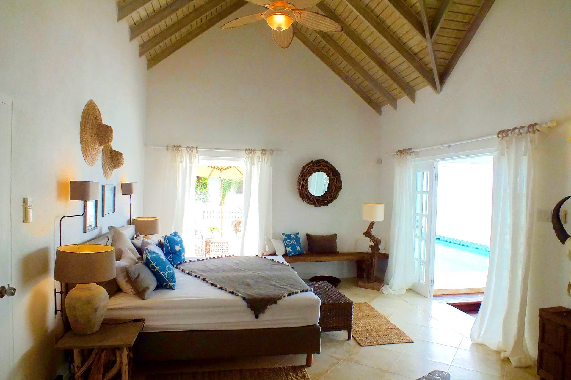 Bright bedroom with vaulted wood ceiling, white walls, bed, blue accents, and open doors to a sunny balcony