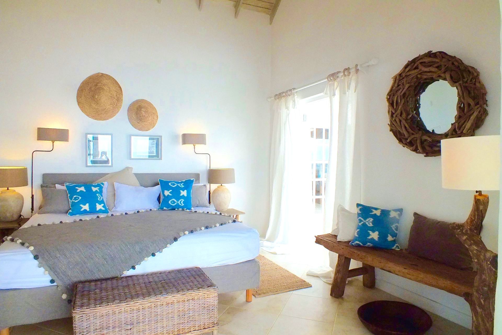 Bright bedroom with a bed, blue pillows, woven wall decor, and a wooden bench by a sunlit window.