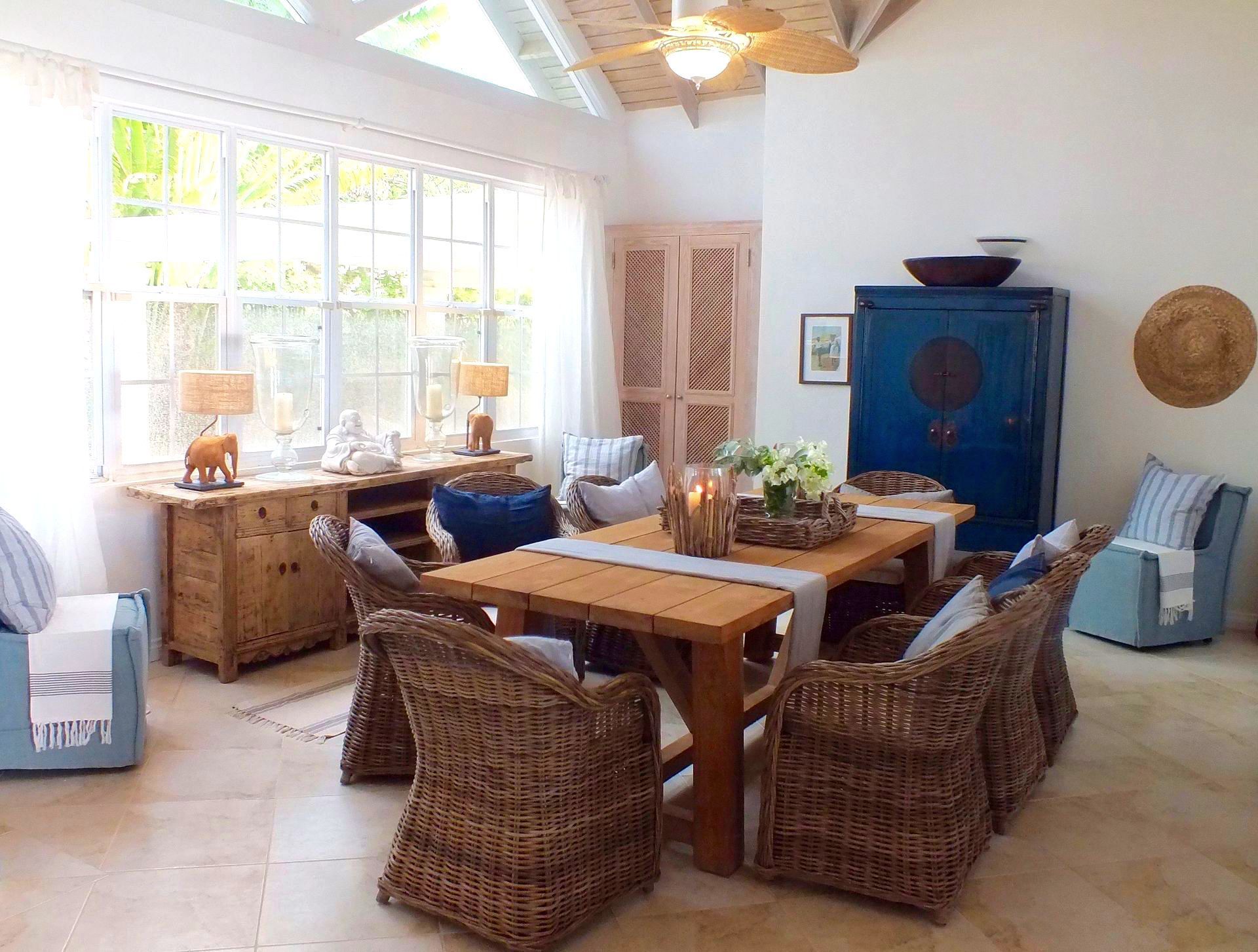 Bright sunroom with wicker chairs, wooden table, blue accents, and large windows overlooking a cozy seating area