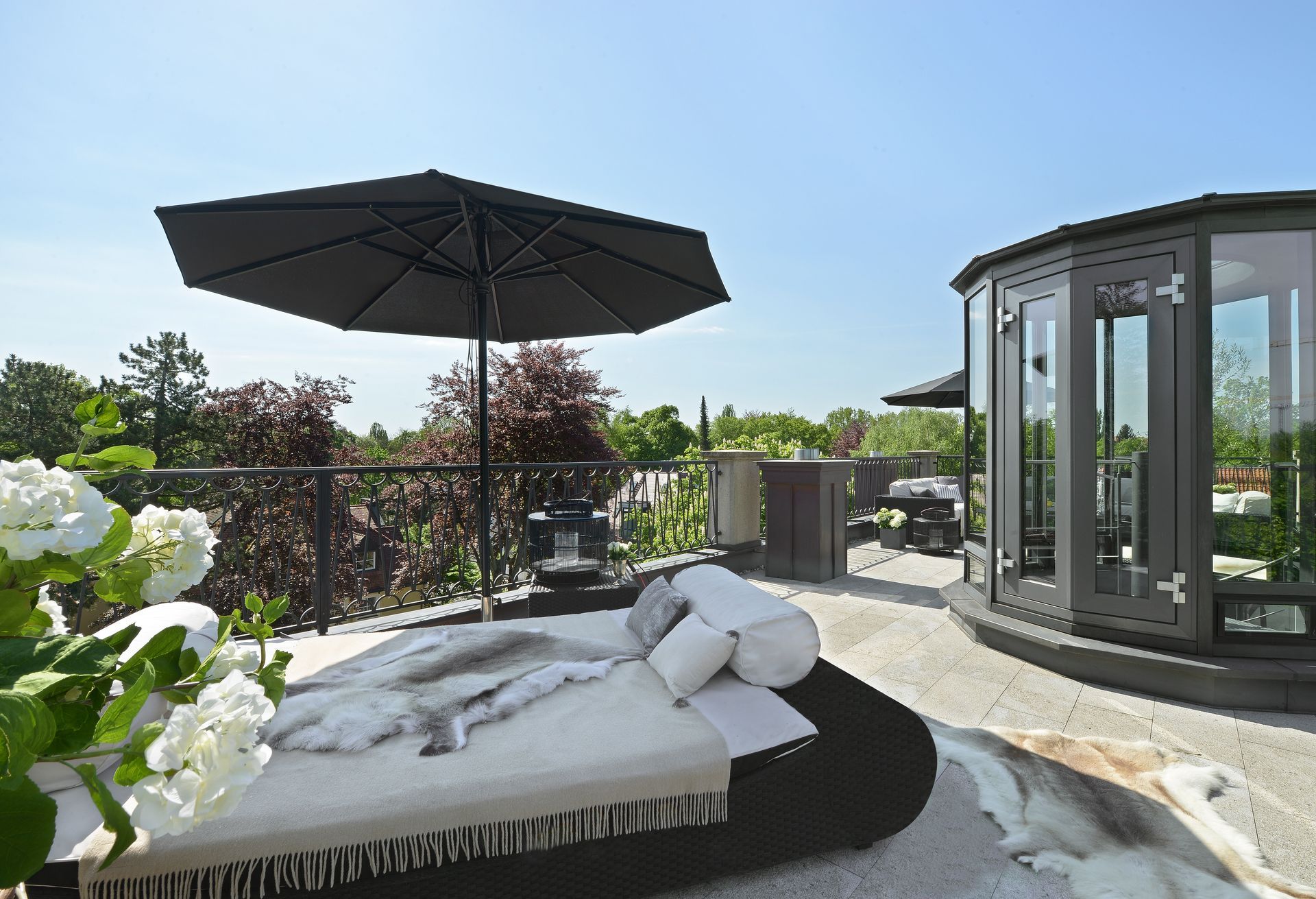 Sunny patio with lounge bed, black umbrella, and glass gazebo beside flowering plants