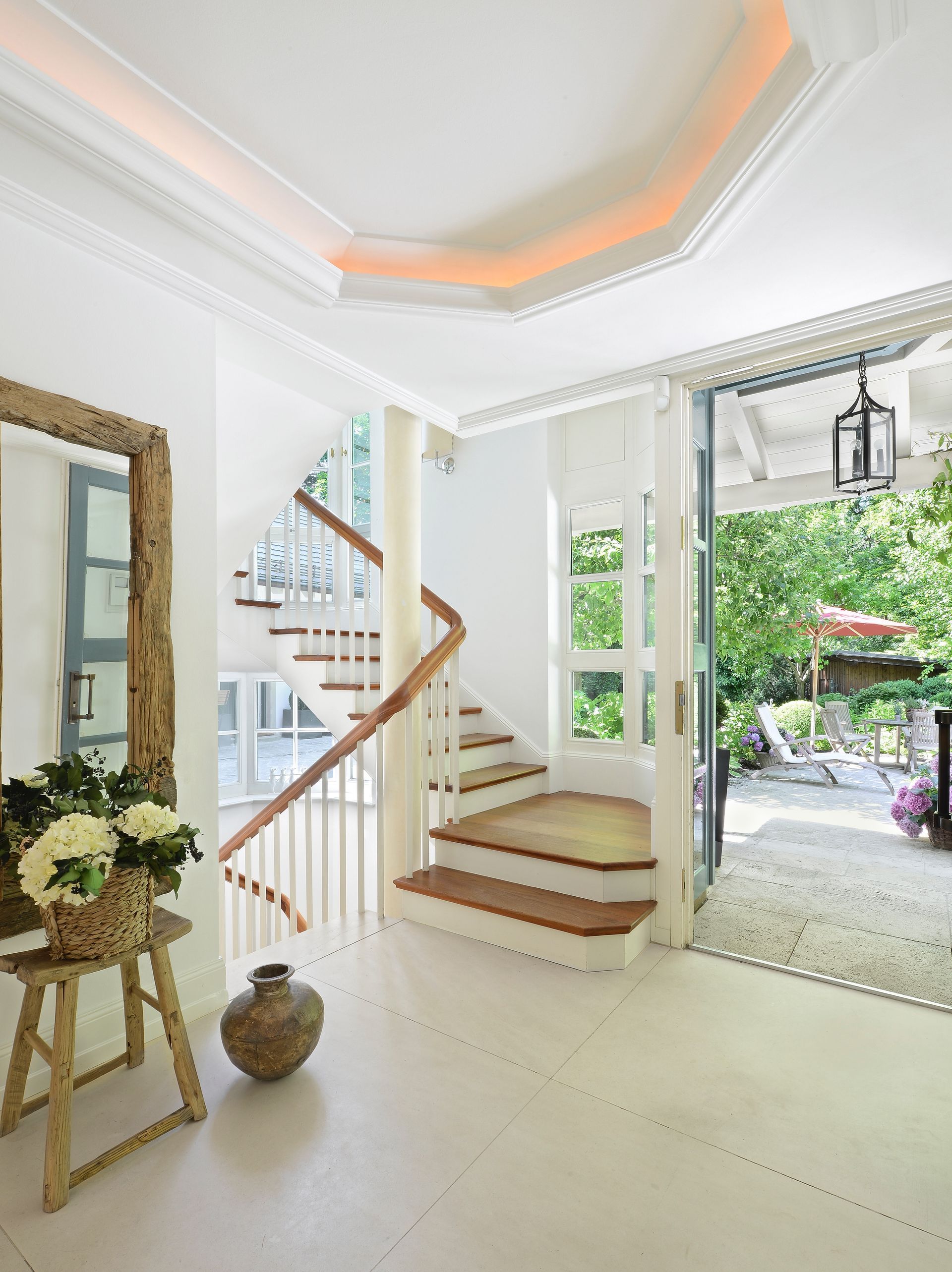Bright white foyer with staircase, mirror, plants, and open doors to a green garden.