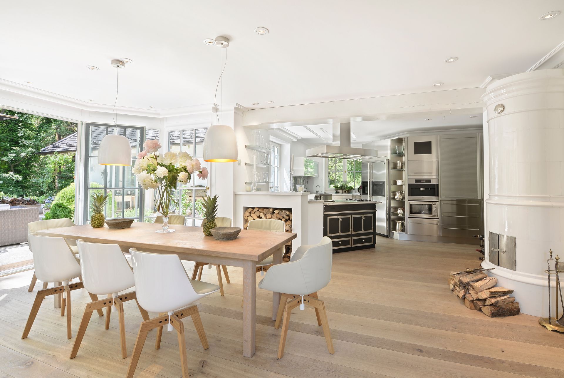 Bright open dining room with wooden table, white chairs, fireplace, and large windows overlooking greenery