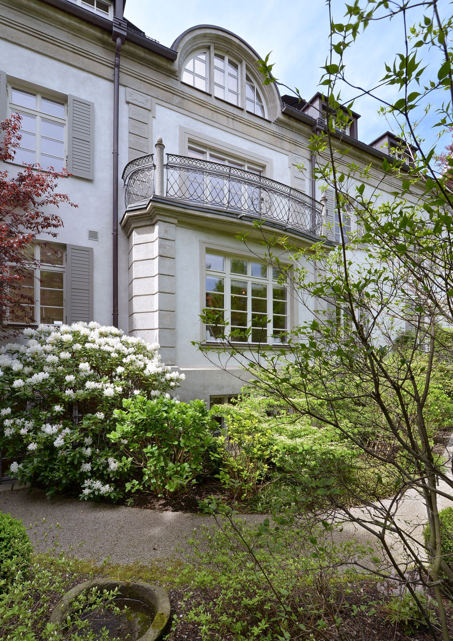 White house facade with arched balcony, large windows, and flowering shrubs in the garden