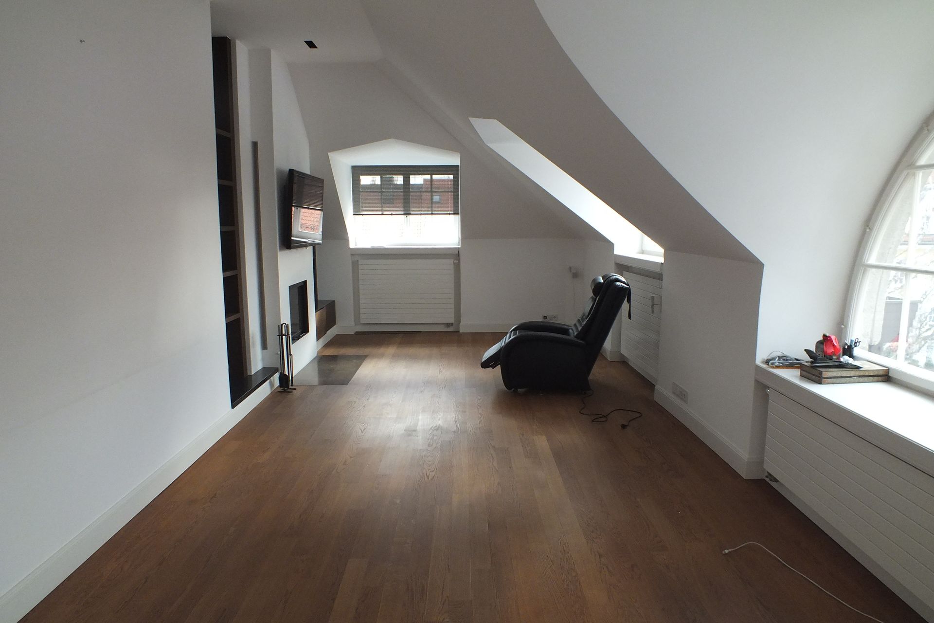 Attic room with wooden floor, white walls, a recliner, and a small window at the far end