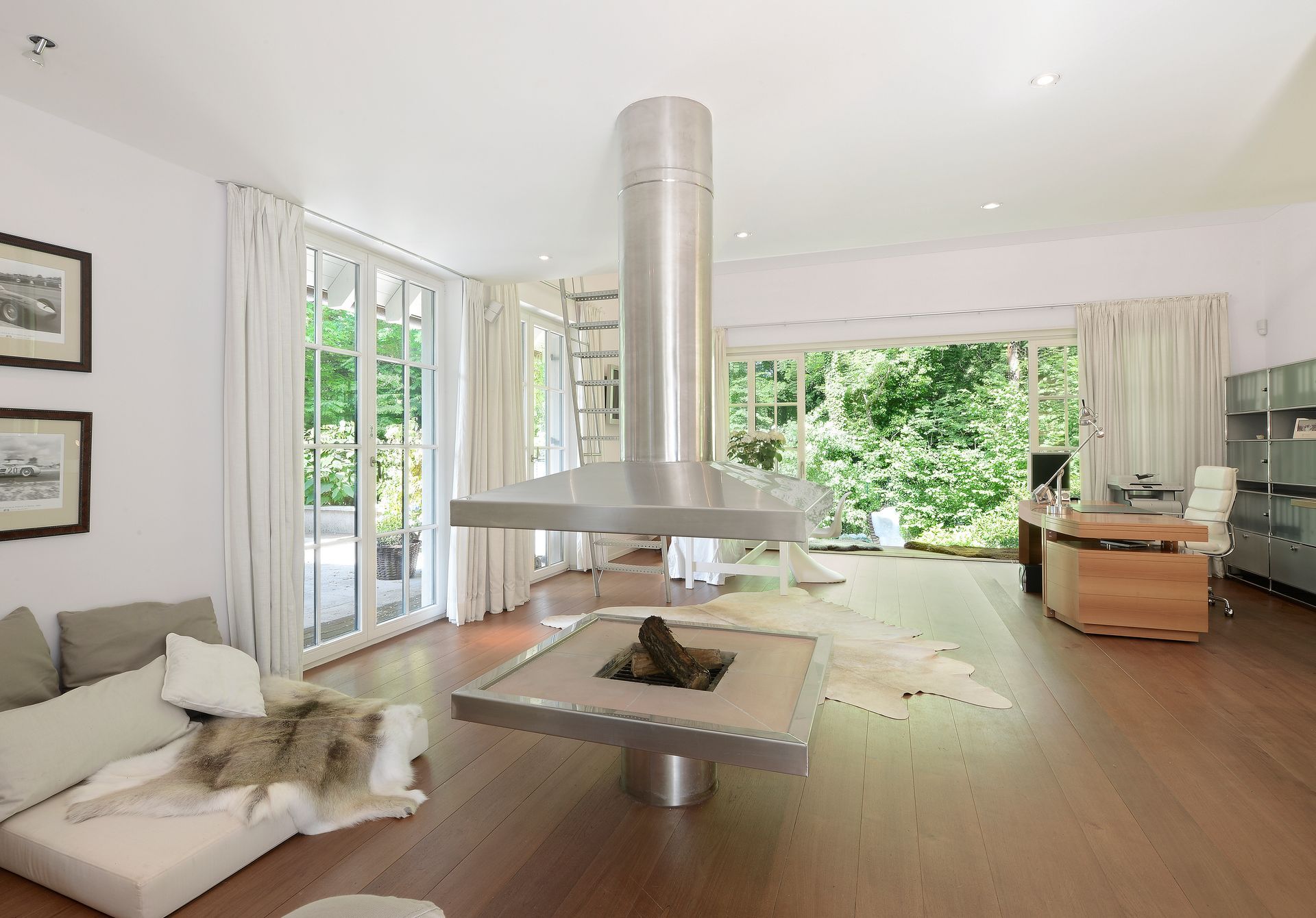 Modern living room with a central fireplace, large windows, white furnishings, and wood floors