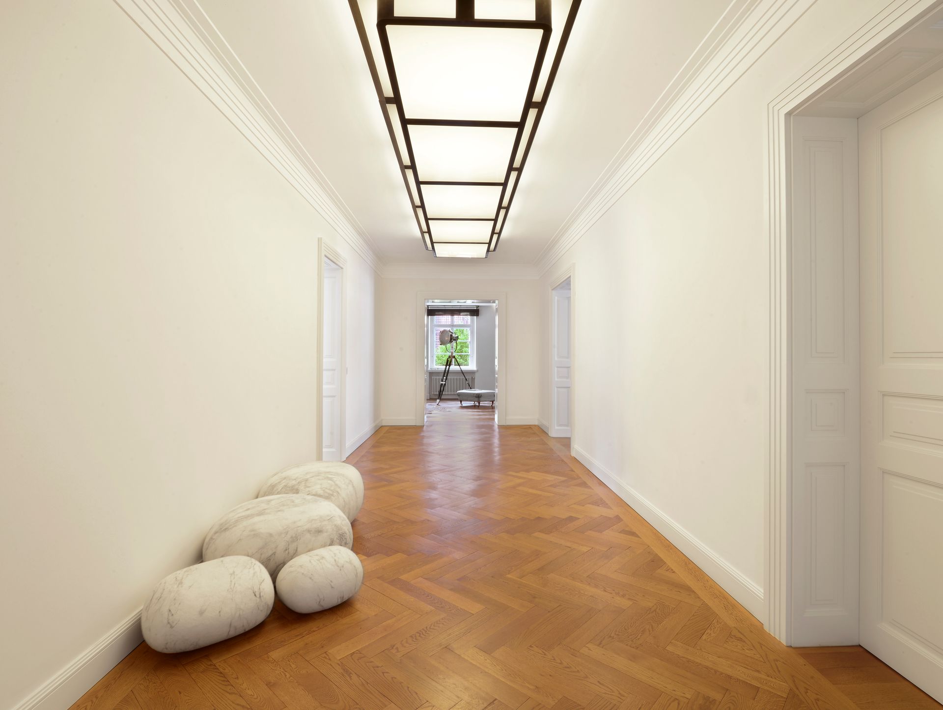 Bright hallway with white walls, herringbone wood floor, plush stools, and a ceiling light panel.