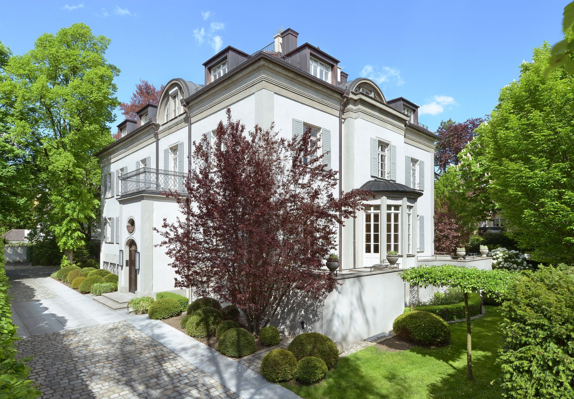 White historic house with ornate trim, red-leaf tree, and a green garden under a blue sky