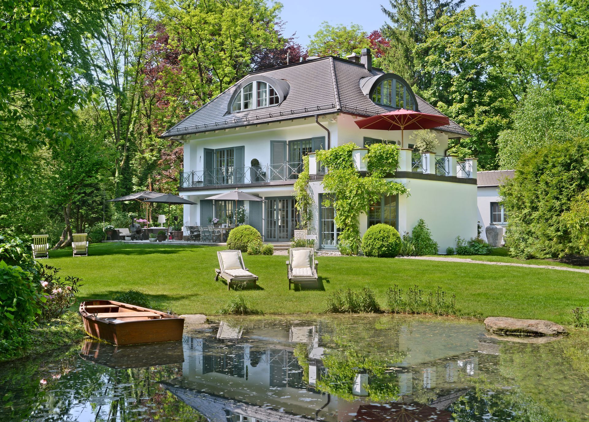 White house with dark roof beside a pond and lush green garden in sunlight