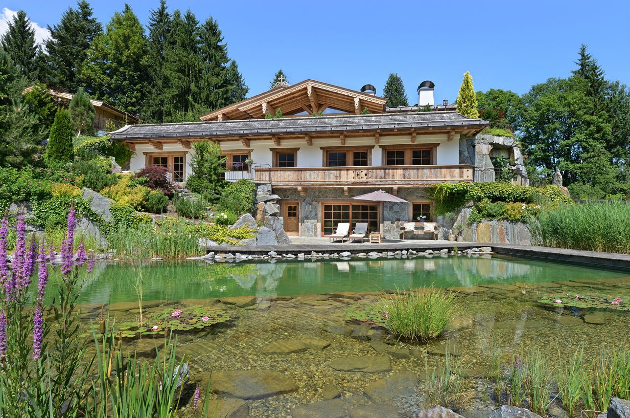 Rustic lakeside villa with a wooden balcony, reflecting pond, and lush green trees under a blue sky