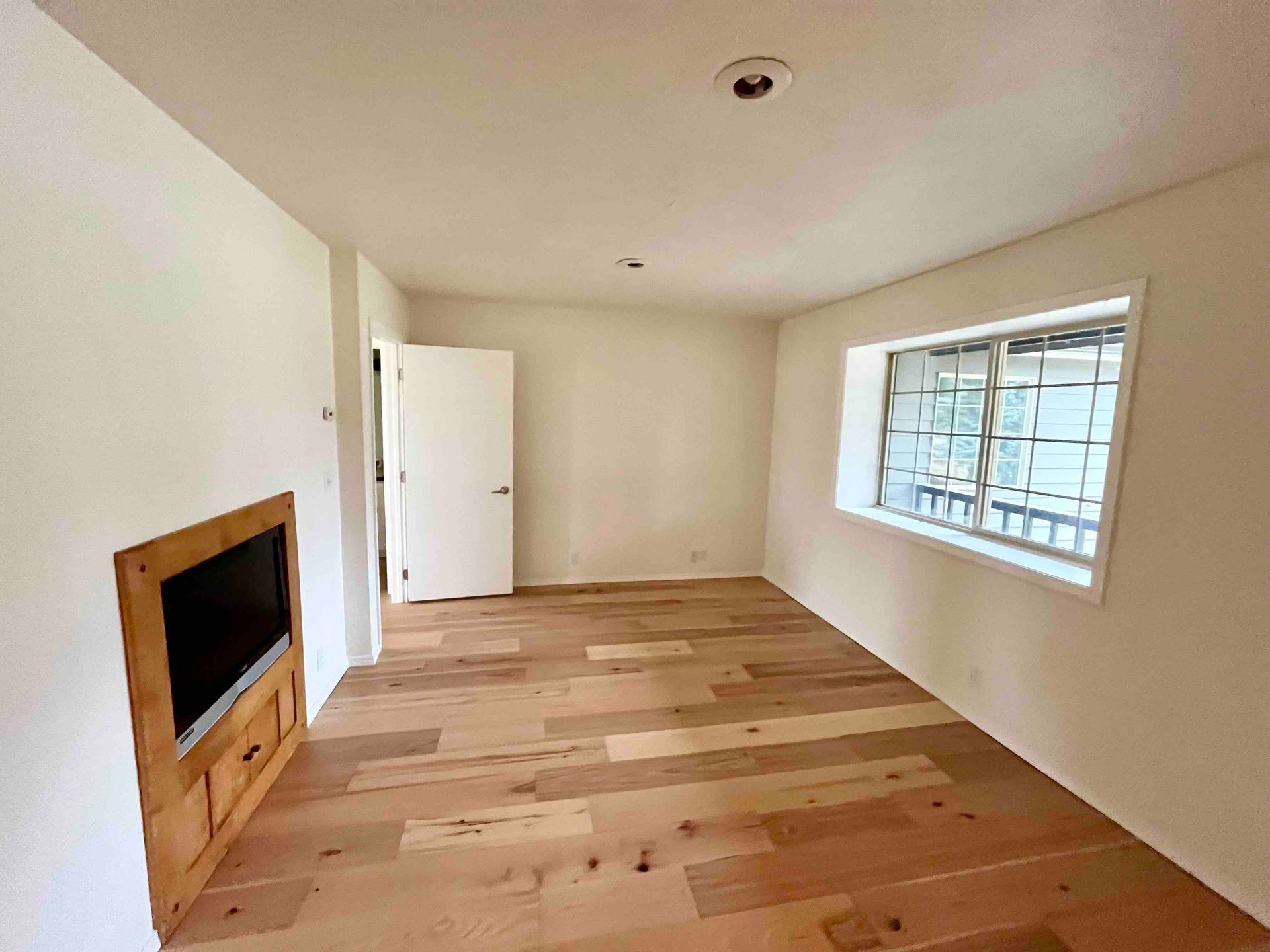 Empty living room with light wood floor, white walls, fireplace, open door, and large window.
