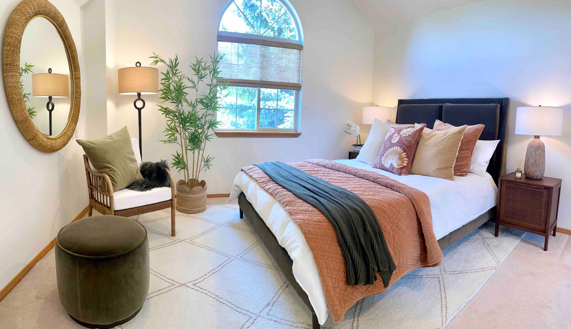 Bright bedroom with a bed, armchair, round mirror, lamp, and arched window in soft neutral tones