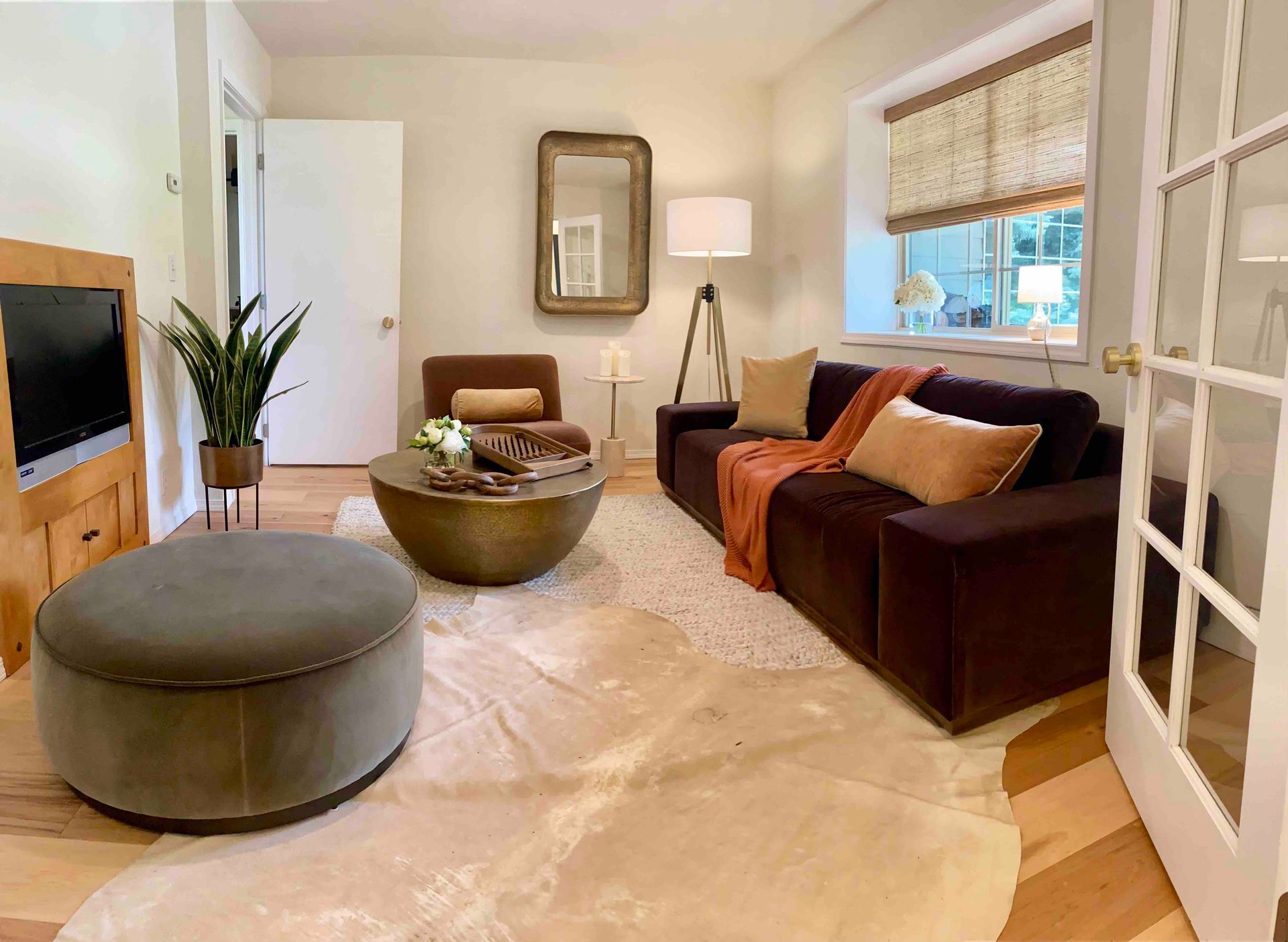 Bright living room with a brown sofa, round ottoman, beige rug, and window with woven shade.