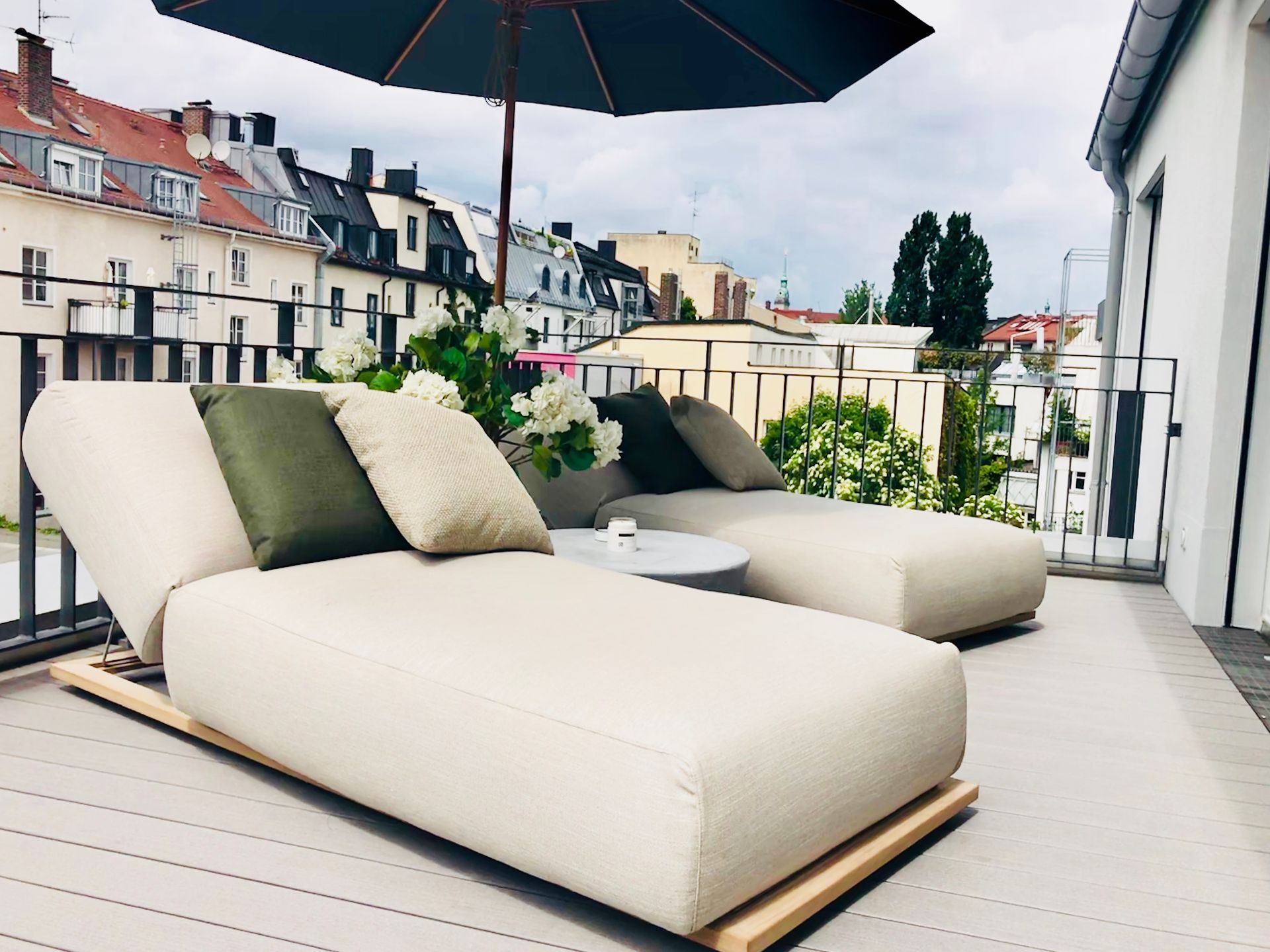 Modern rooftop terrace with cream chaise lounges, green pillows, and a large umbrella overlooking city buildings