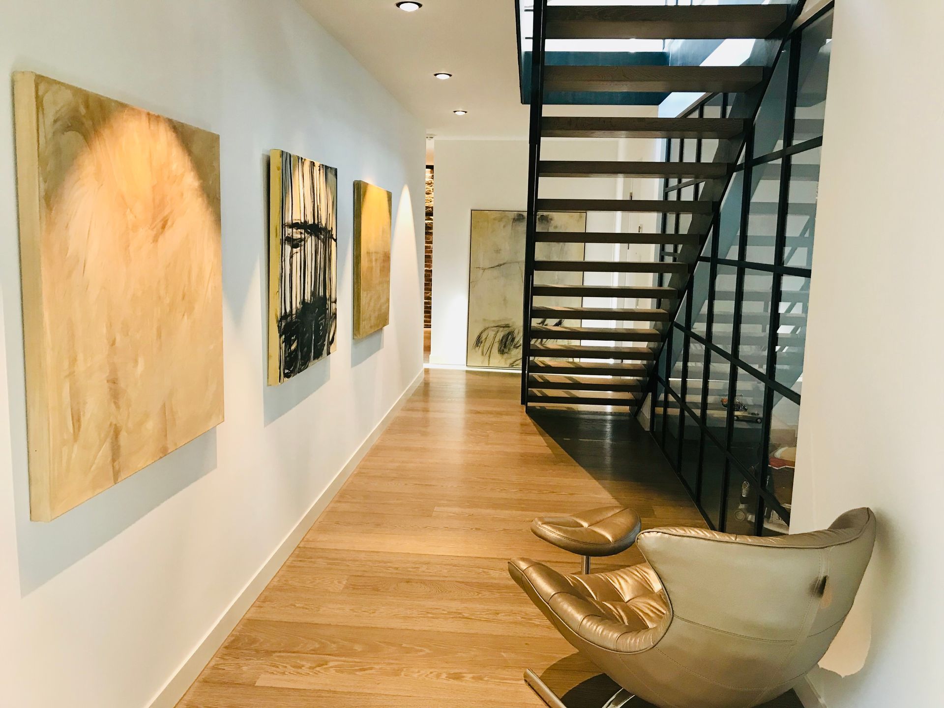 Modern hallway gallery with abstract art, wood floors, black staircase, and a sculptural chair