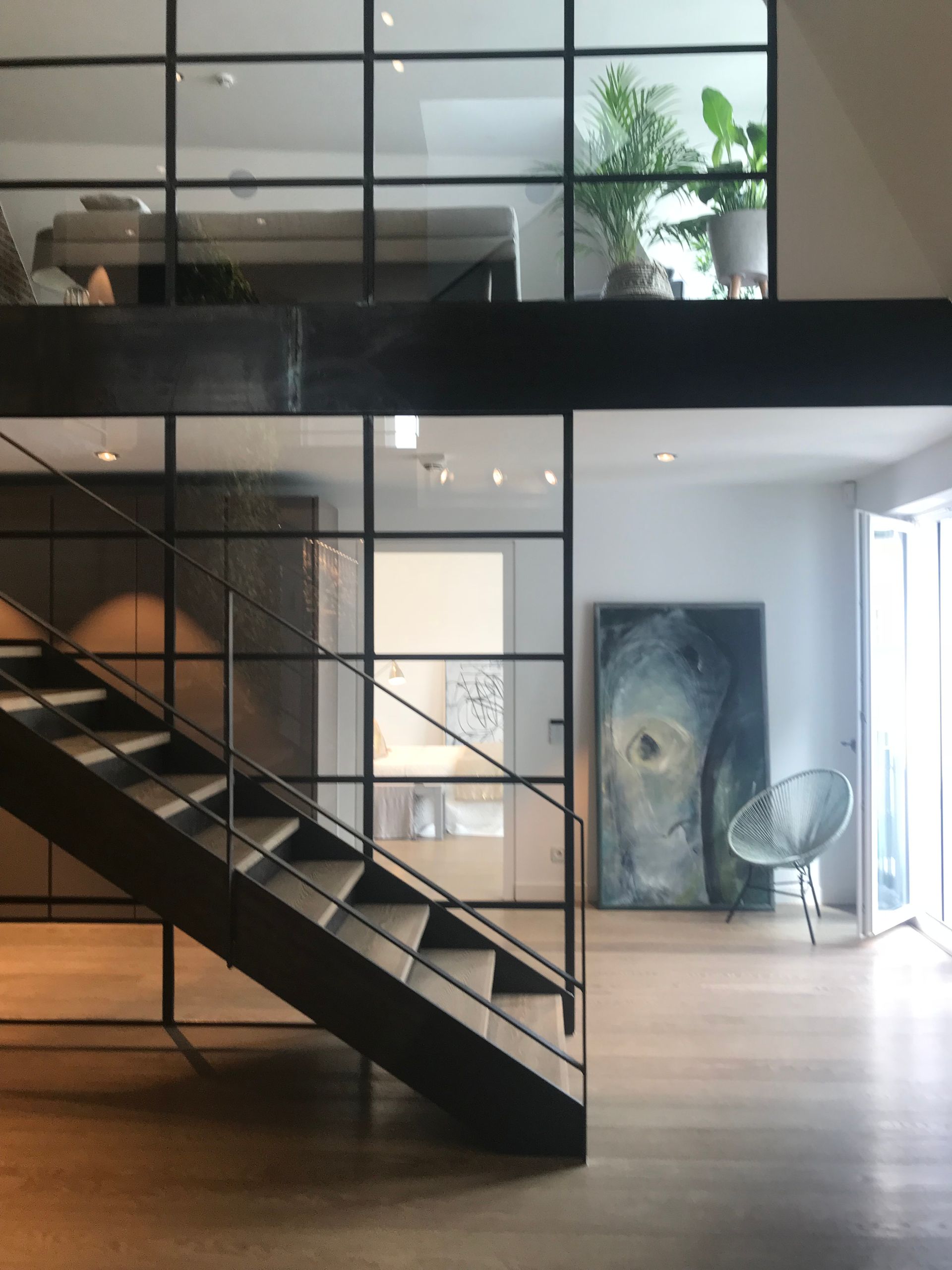 Modern loft interior with black metal staircase, glass walls, and a chair beside abstract art.