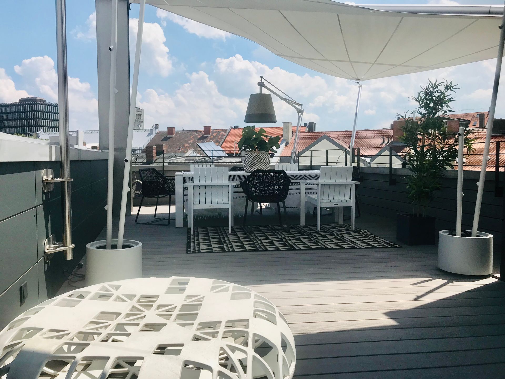 Rooftop terrace with white tables and chairs under a canopy, overlooking city rooftops and a blue sky.